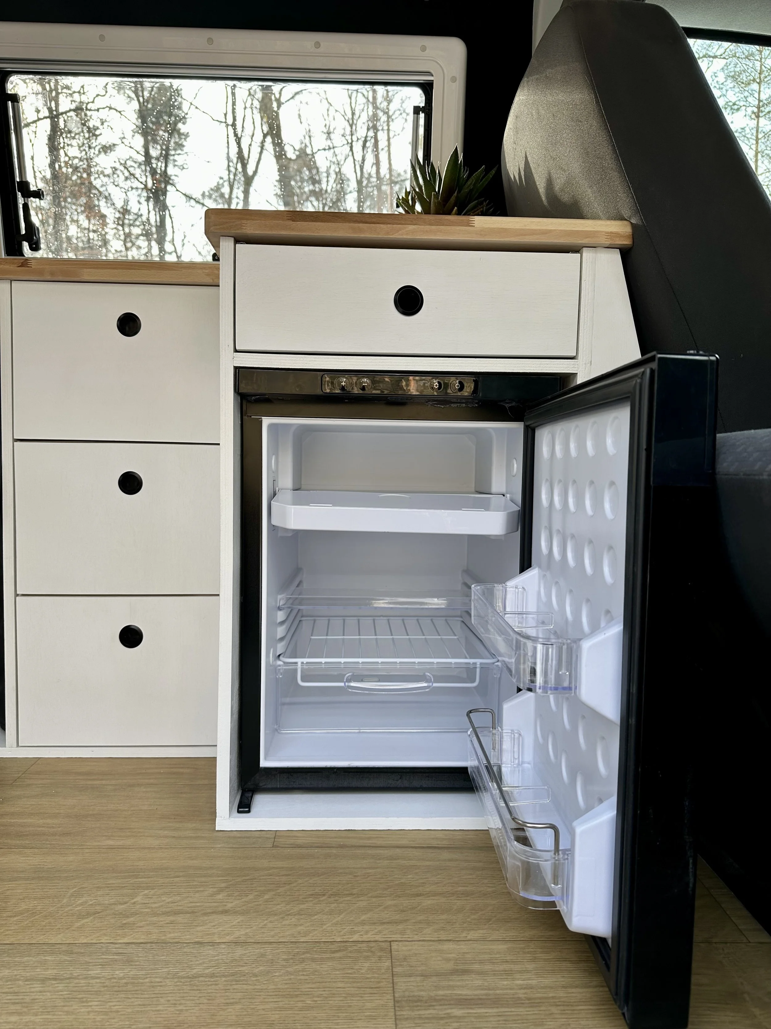 Ein kleiner, offener Kühlschrank in einer Wohnmobilküche, mit leeren Regalen und transparenten Ablagefächern, neben einem weißen Schrank mit weißen Schubladen und einem Holzarbeitsplatte. Im Hintergrund sind Fenster mit Blick auf Bäume im Freien.