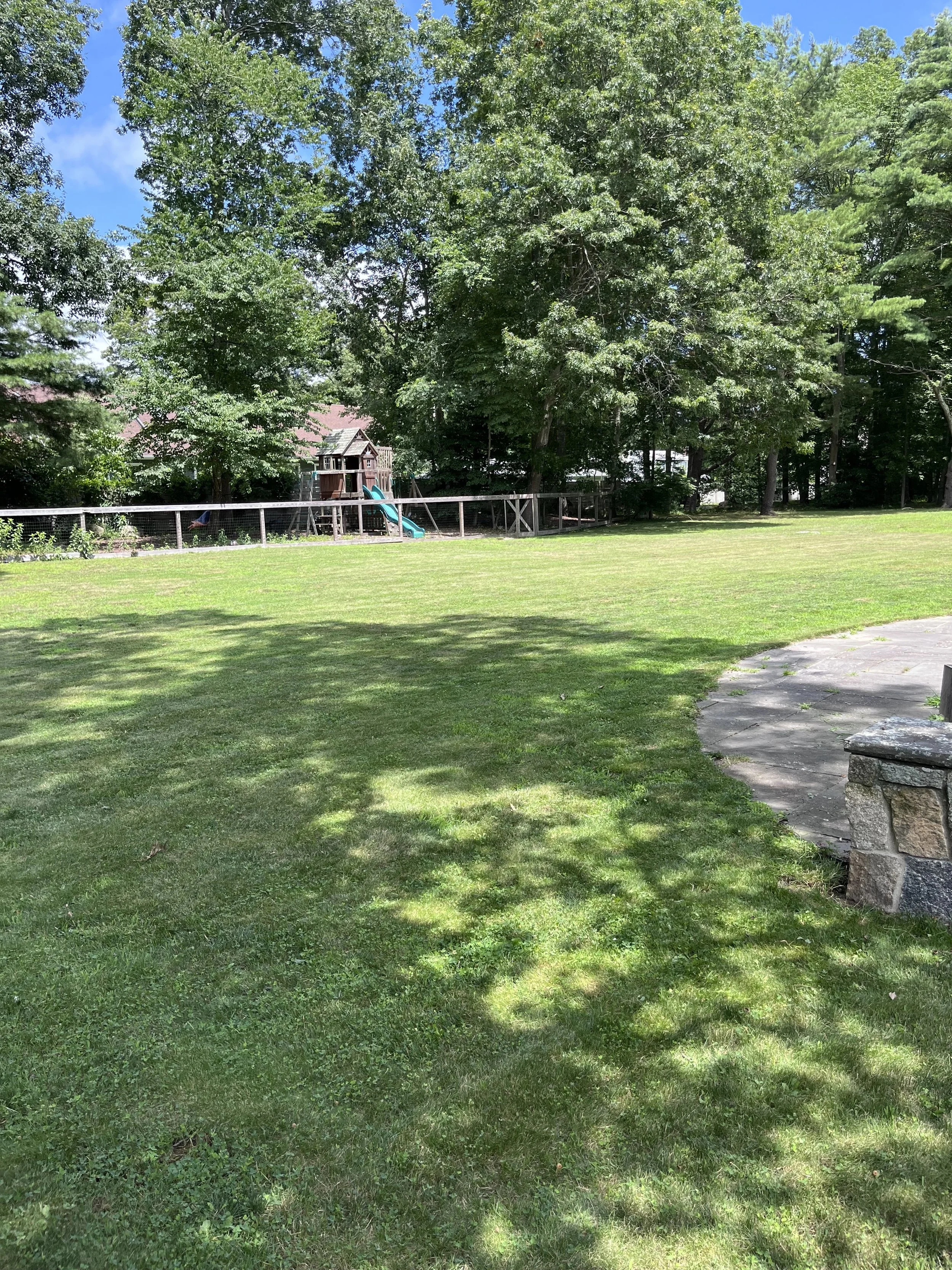 A backyard with a freshly cut lawn, a clean concrete walkway, a stone bench, and a wooden children's play structure with a slide, surrounded by trees.
