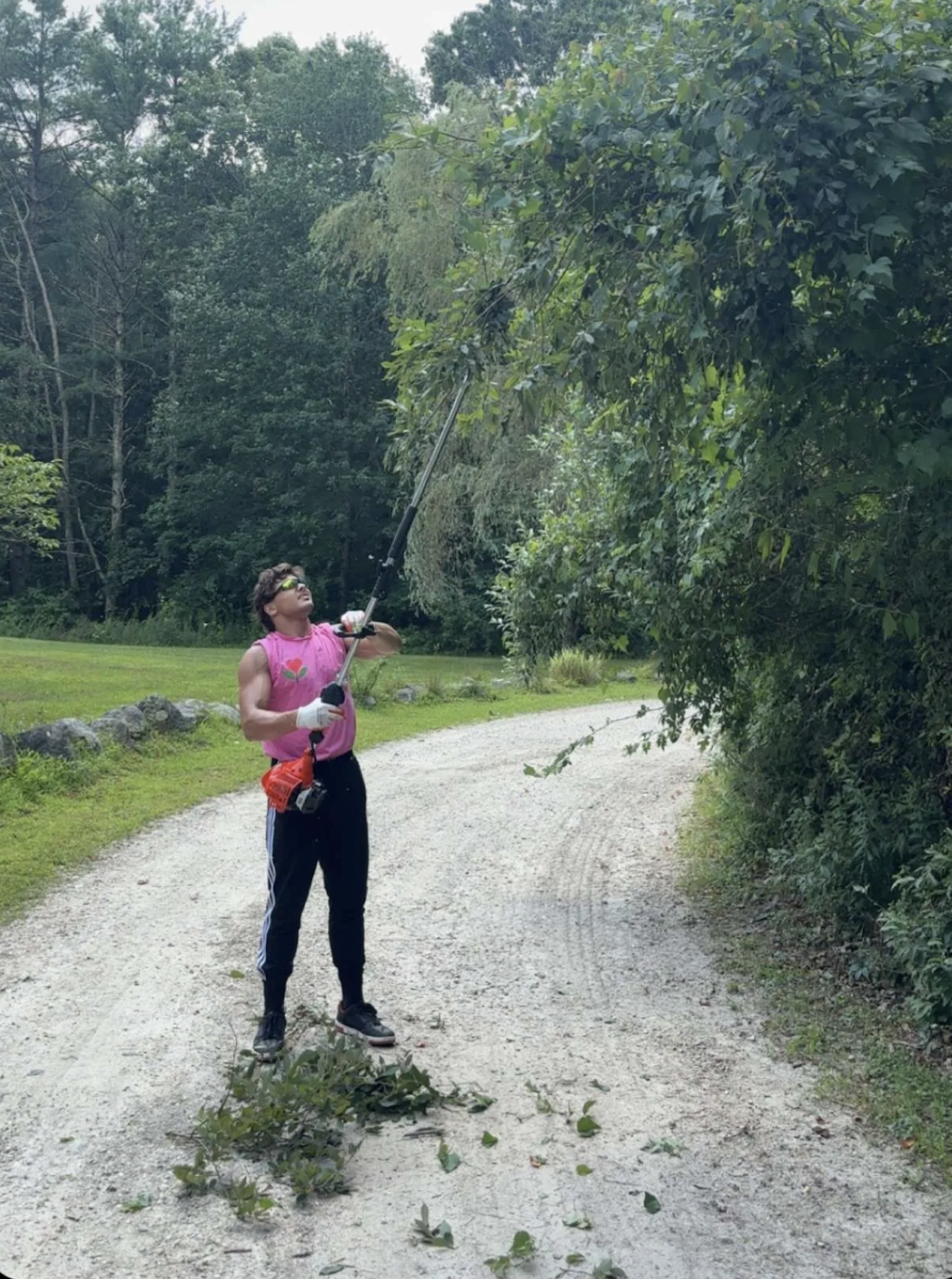 Matt Ingalls standing on a gravel road and trimming branches from a tree with a pole saw. He is wearing sunglasses, a pink sleeveless shirt, black track pants, and gloves, with a red tool bag hanging from their waist.