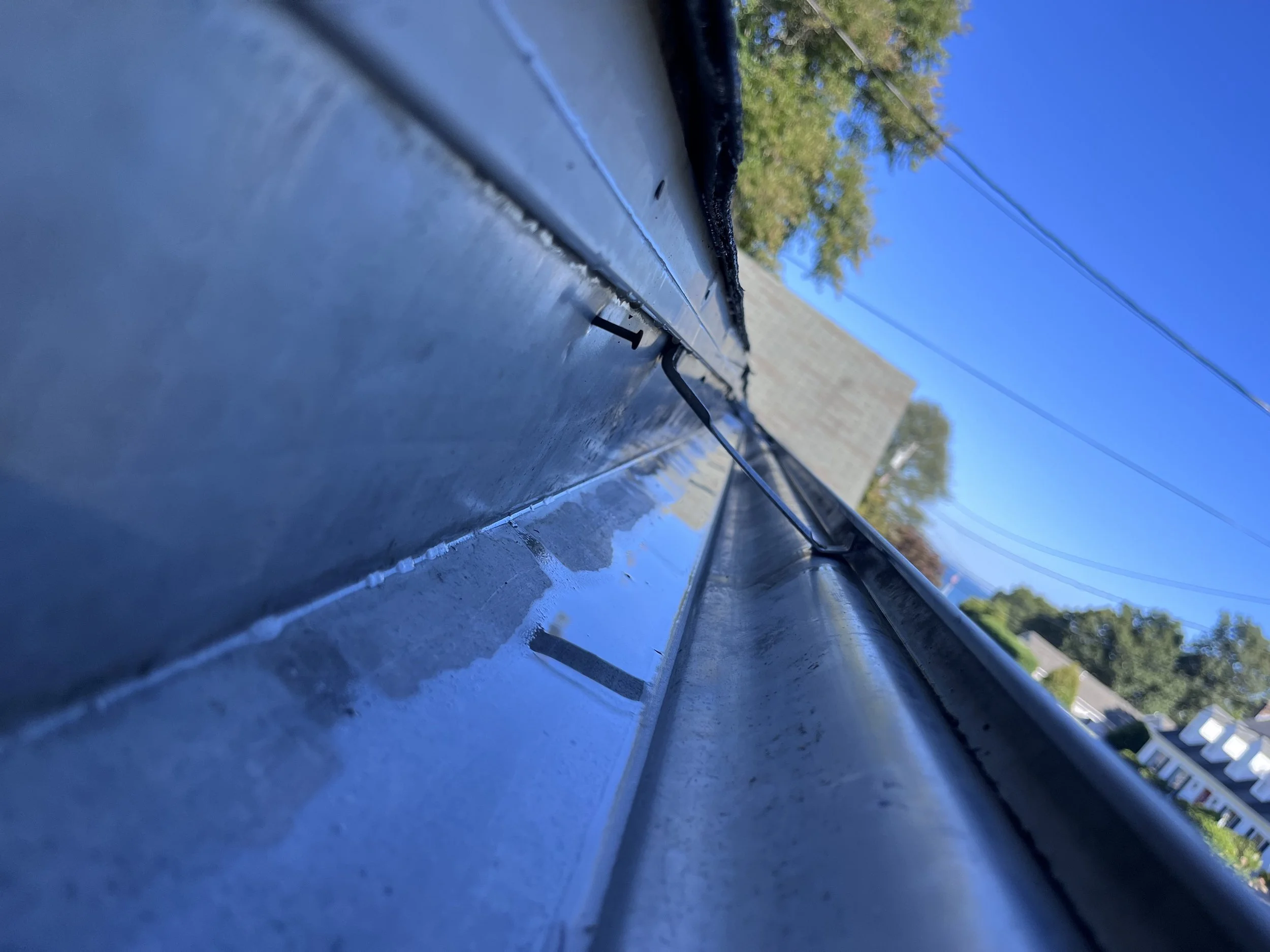 Close-up image of a clean, shiny metal gutter with a roof vent, with a bright blue sky and house rooftops in the background.