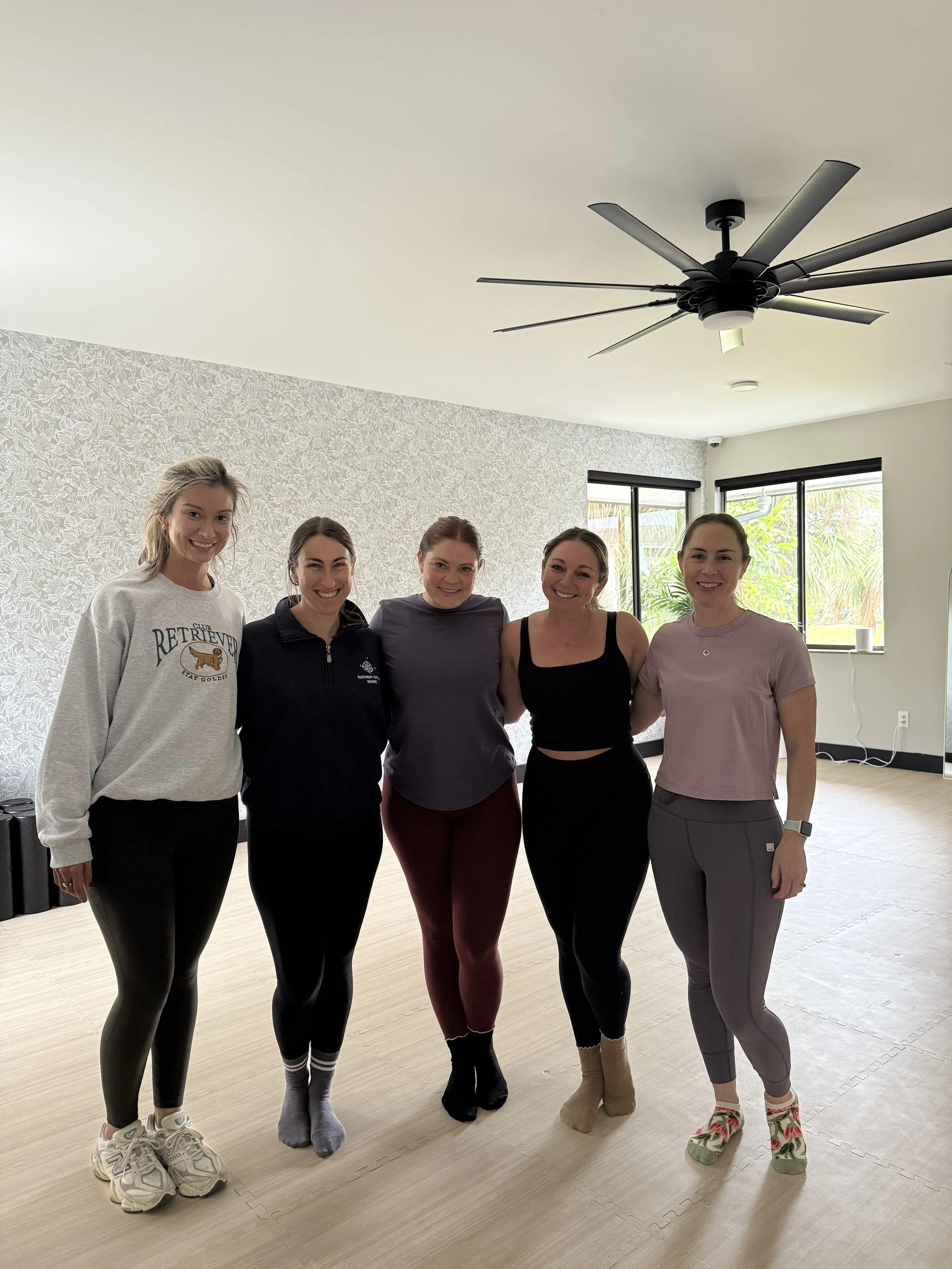 Five women standing together in a bright room with large windows and a ceiling fan, smiling at the camera.