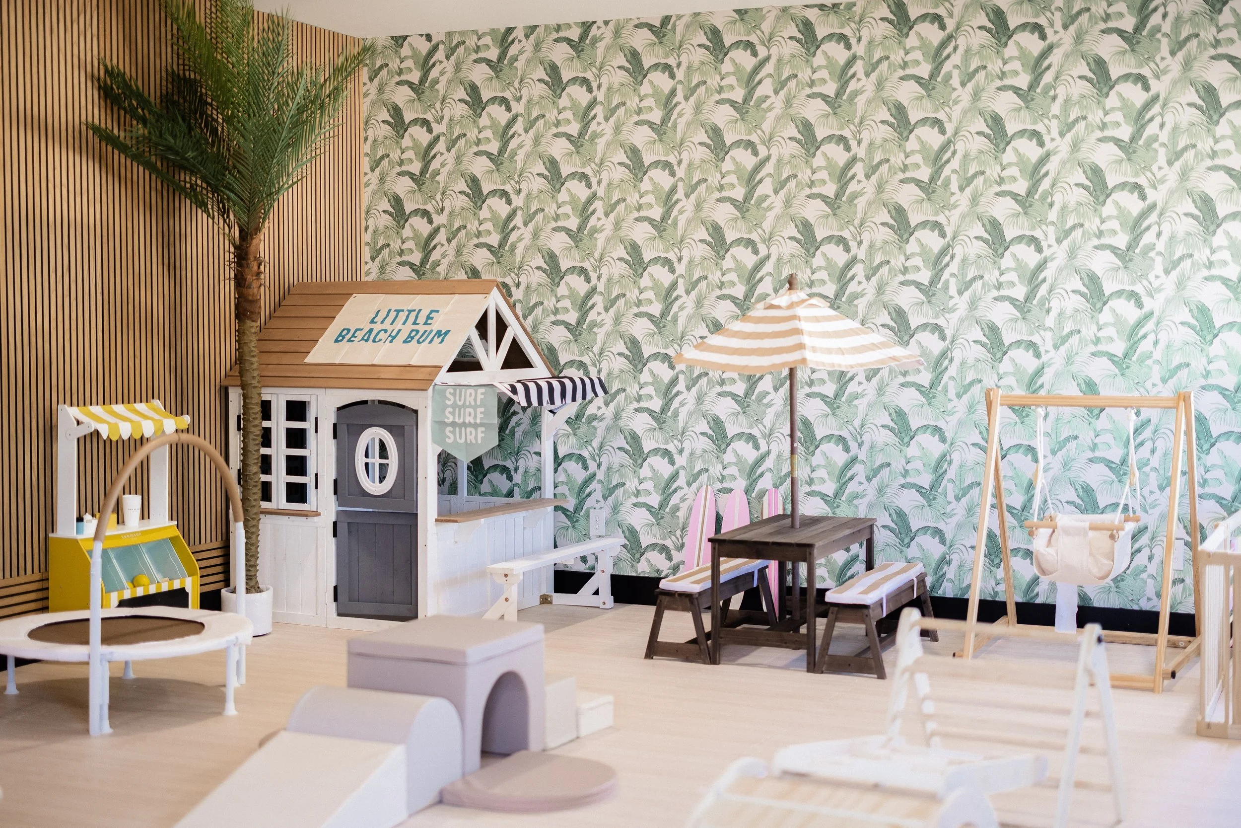 Indoor play area with beach-themed decor, including a small playhouse labeled 'Little Beach Bum,' a palm tree, a striped umbrella over a wooden table with benches, and a swing with a white pouch, against a wall with tropical leaf wallpaper.