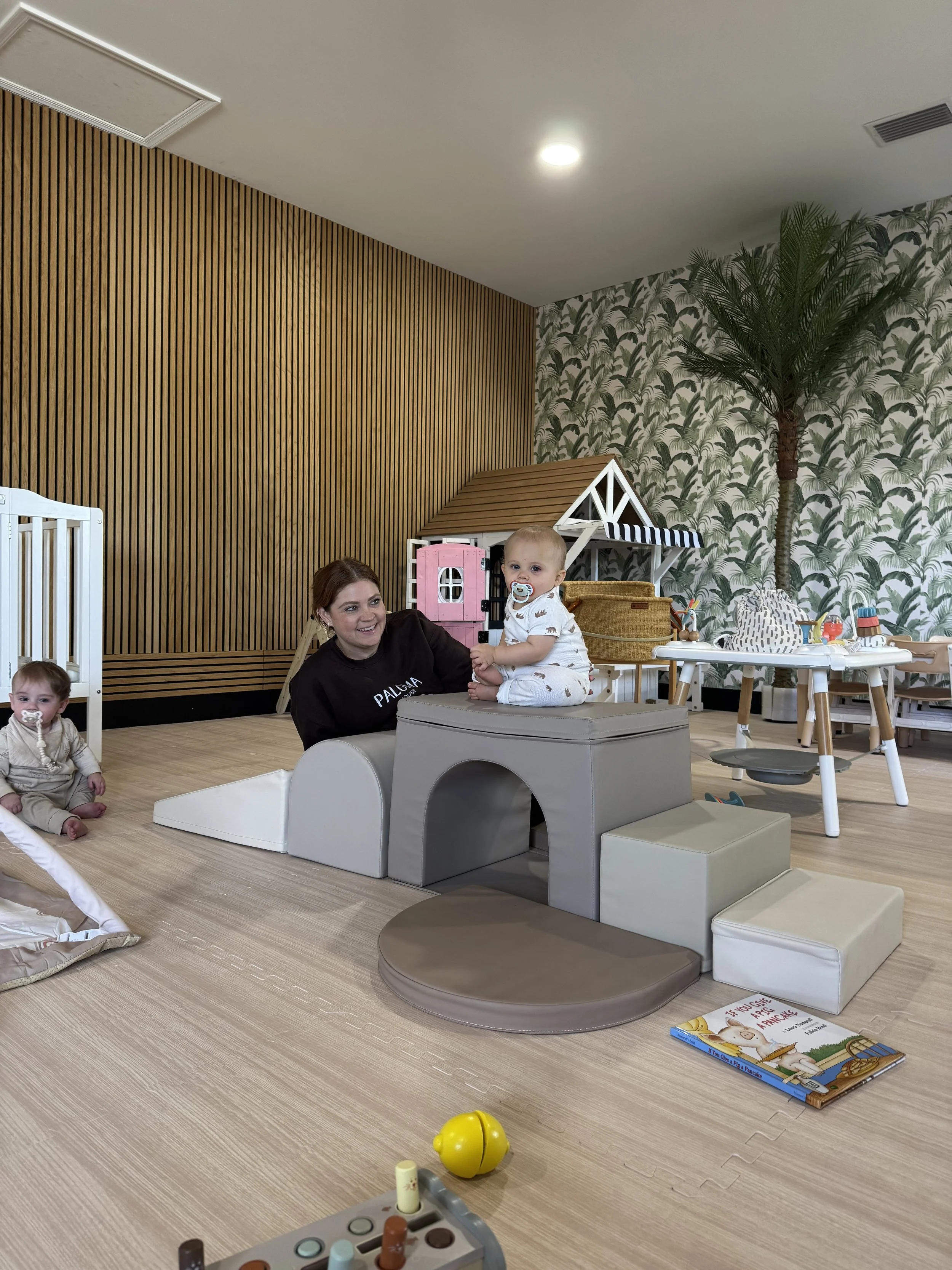 Children playing in a playroom with a woman. Two toddlers are seated on soft play equipment, one with a pacifier. The room features a dollhouse, a round table with toys, a palm tree wallpaper accent wall, and a wooden panel wall.