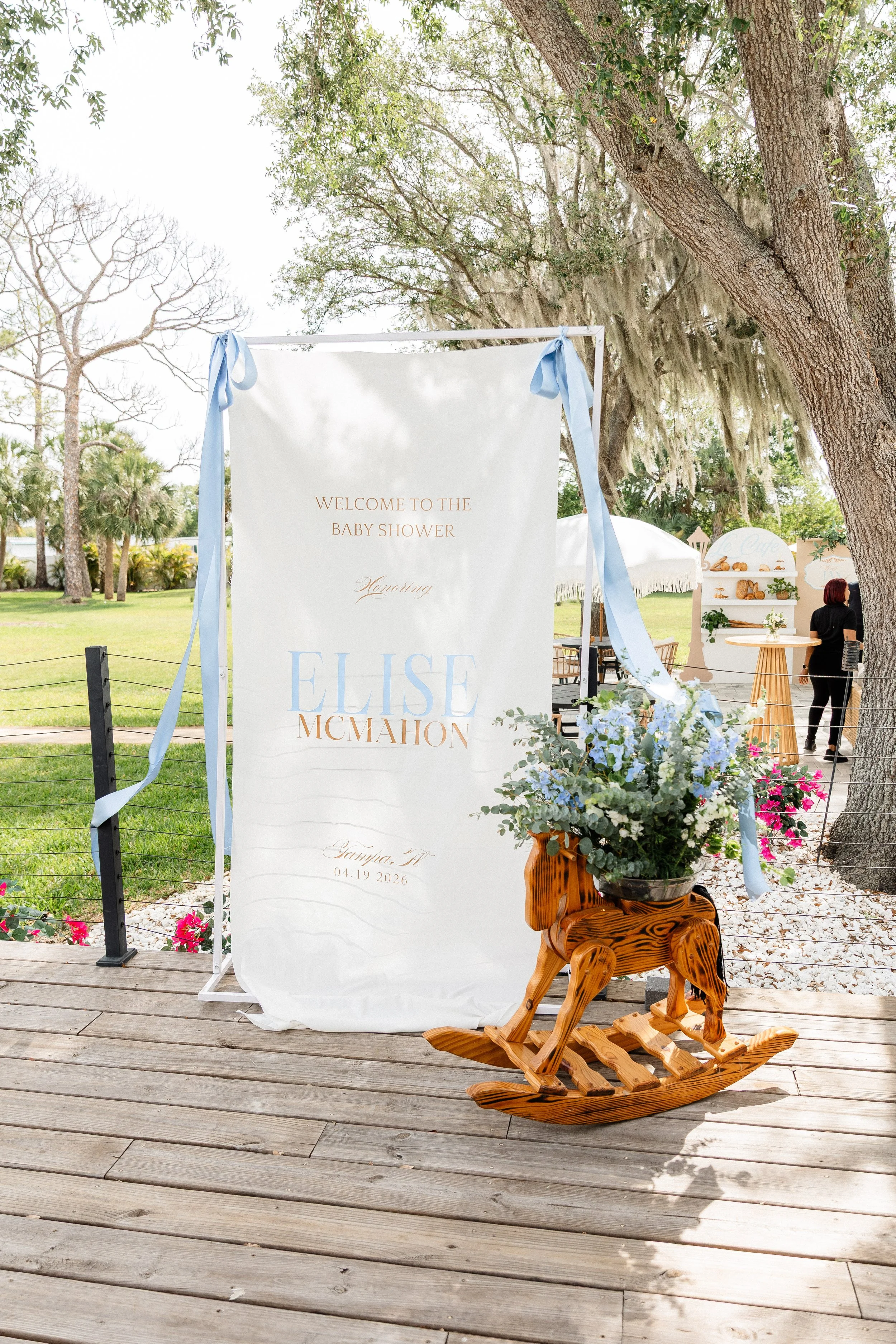 Outdoor baby shower setup featuring a white banner with gold lettering, floral arrangements in a wooden animal-shaped container, and a wooden rocking horse on a wooden deck, surrounded by trees and greenery.
