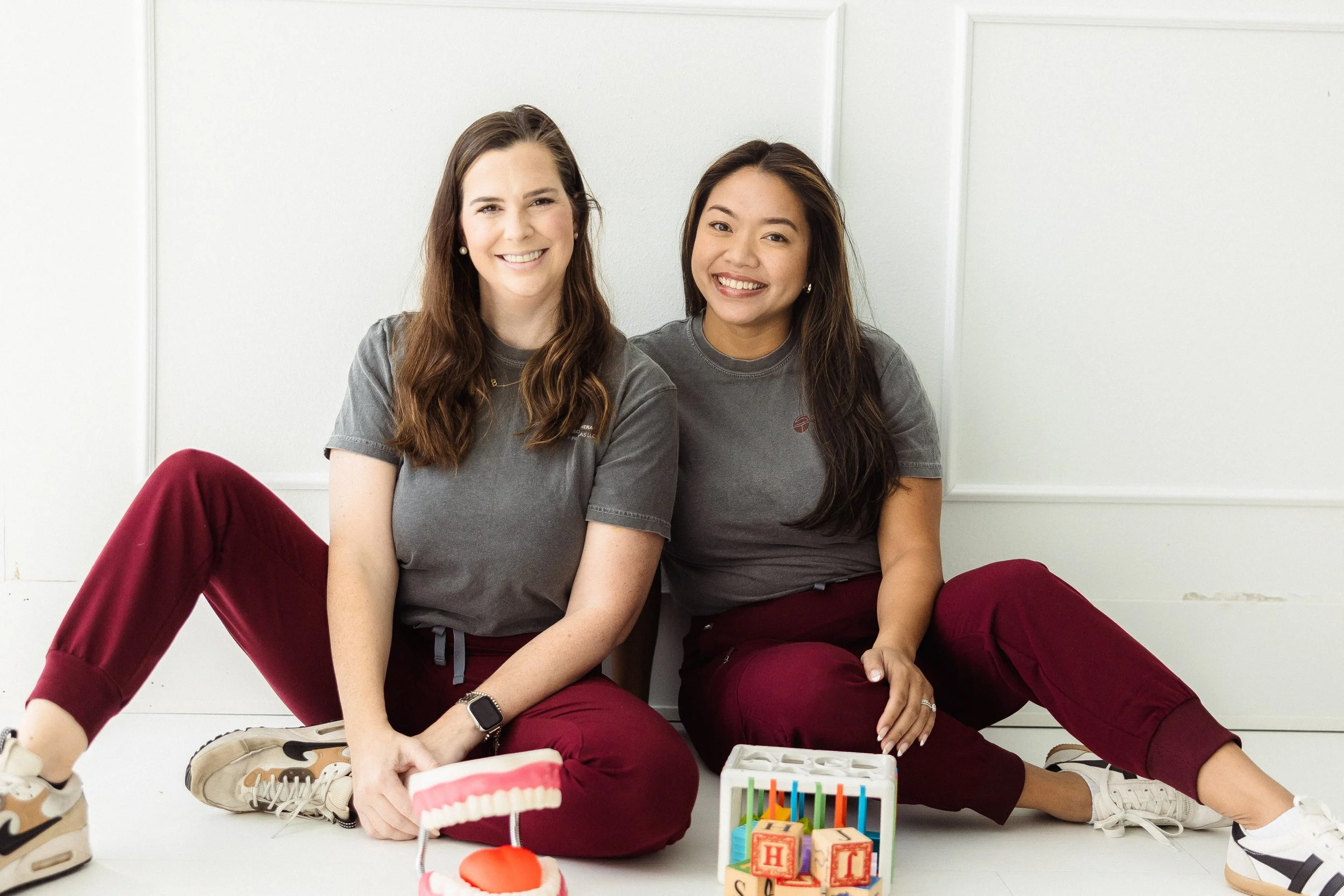 Two women sitting on the floor against a white wall, smiling, surrounded by birthday decorations including a cake with a single candle, a box of colorful blocks with letters, and a red heart-shaped object.