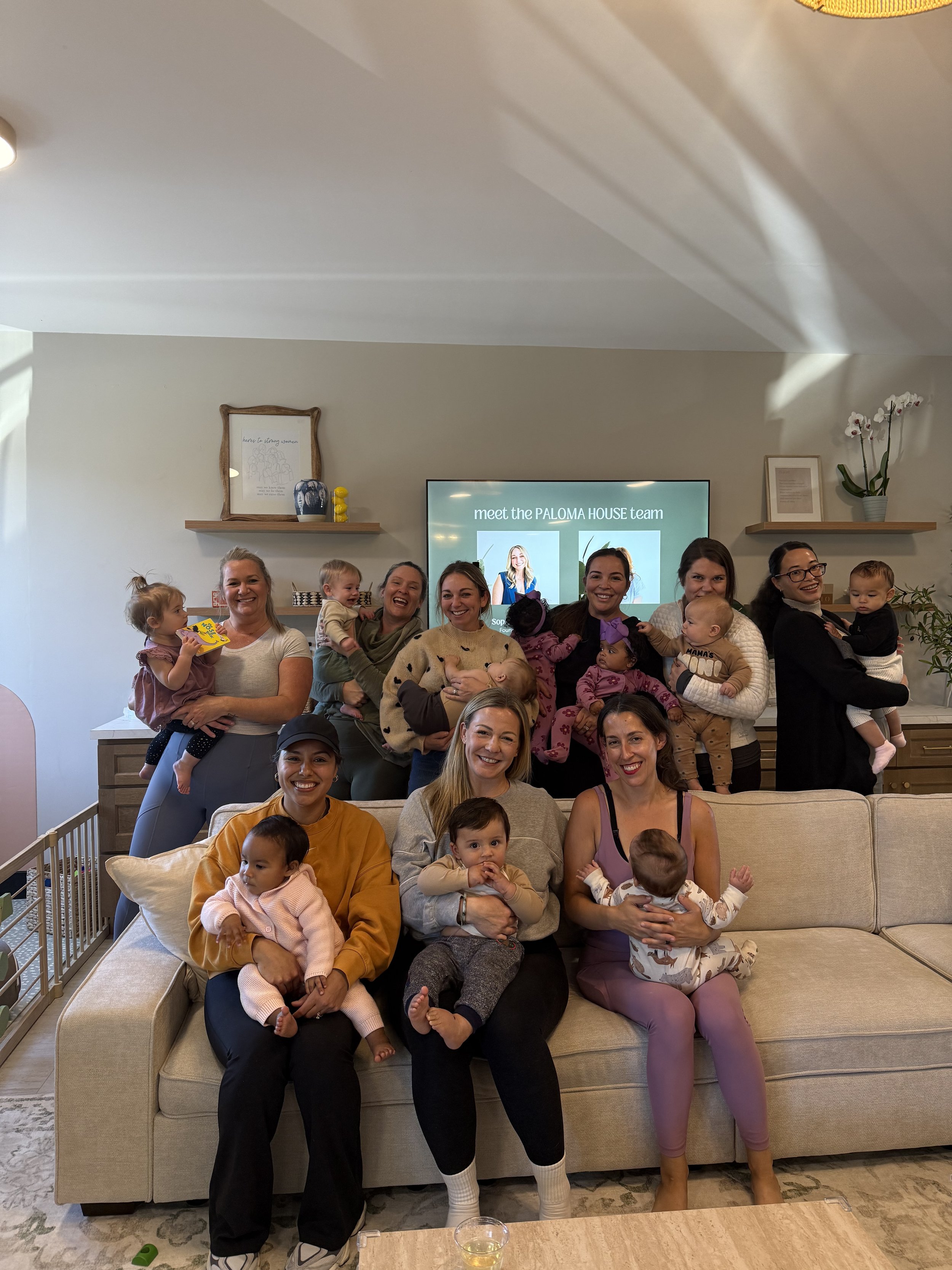 A group of women and children gather in a living room, smiling for a group photo in front of a TV displaying 'meet the PALOMA HOUSE team'. Some women are holding babies, and the children are sitting on their laps or being held. The space is decorated with shelves, framed art, and potted plants.