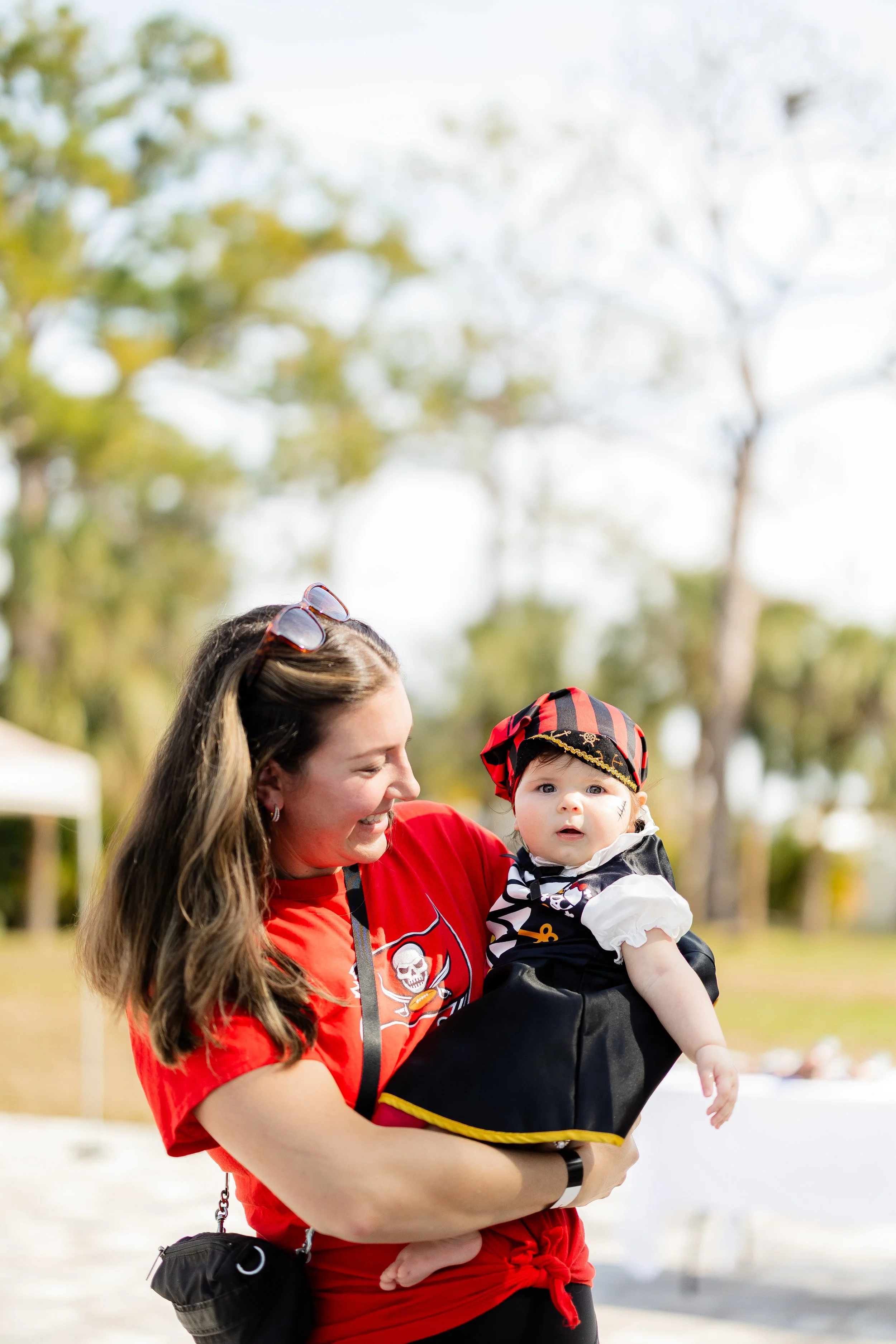 A woman in a red Tampa Bay Buccaneers shirt holding a baby dressed as a pirate outdoors.