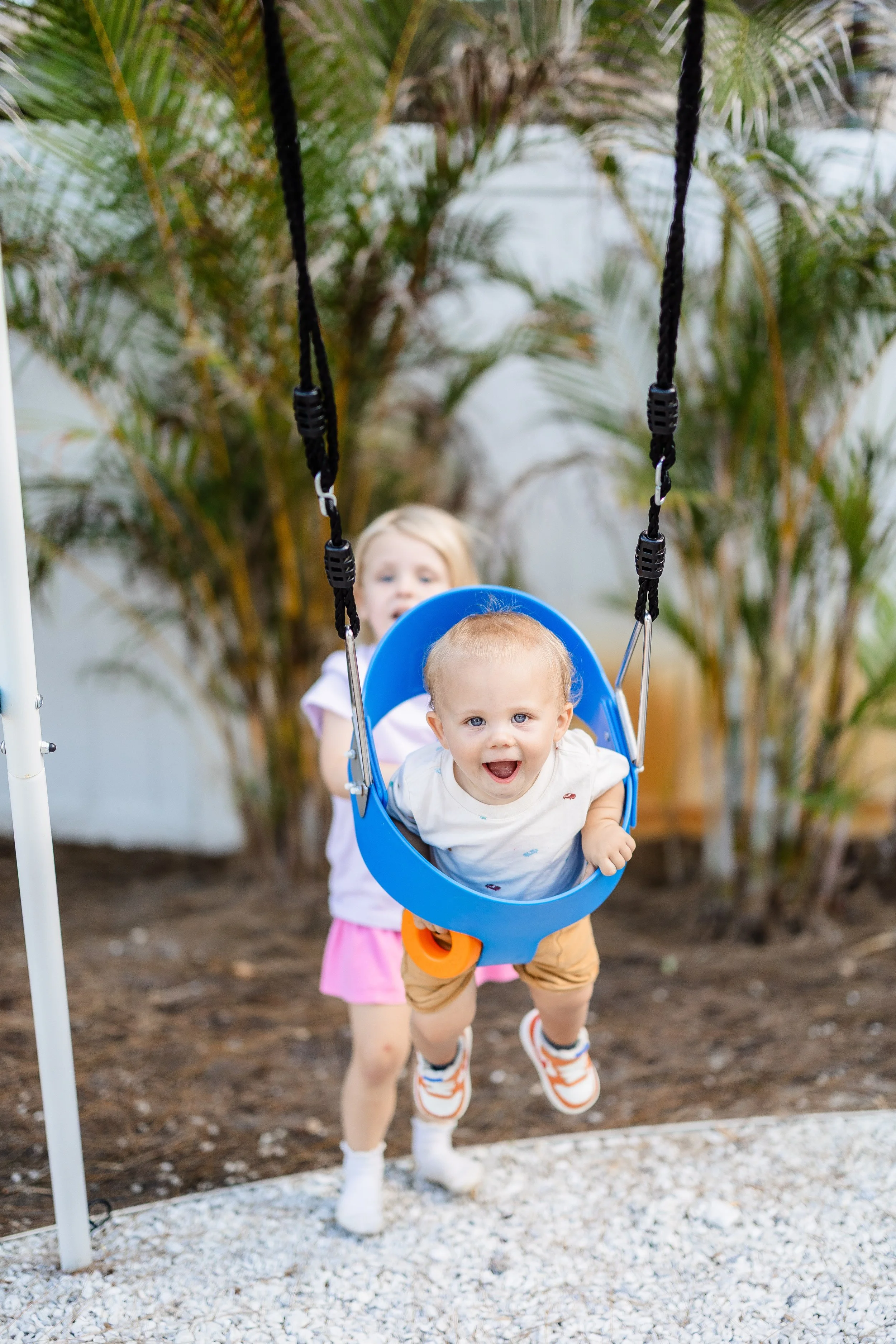Two children playing with a blue baby swing outdoors, surrounded by green plants, with one child sitting in the swing and the other standing nearby, smiling and looking happy.