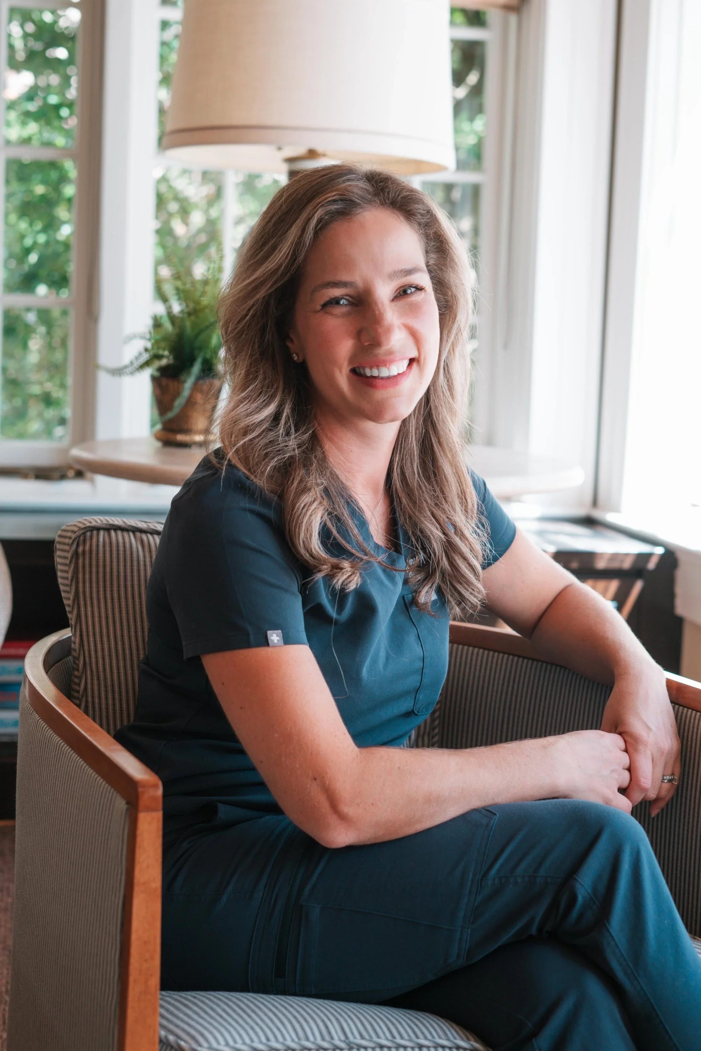 A woman with shoulder-length light brown hair, smiling, wearing dark medical scrubs, sitting on a striped armchair in a well-lit room with large windows, a table lamp, and a potted plant in the background.