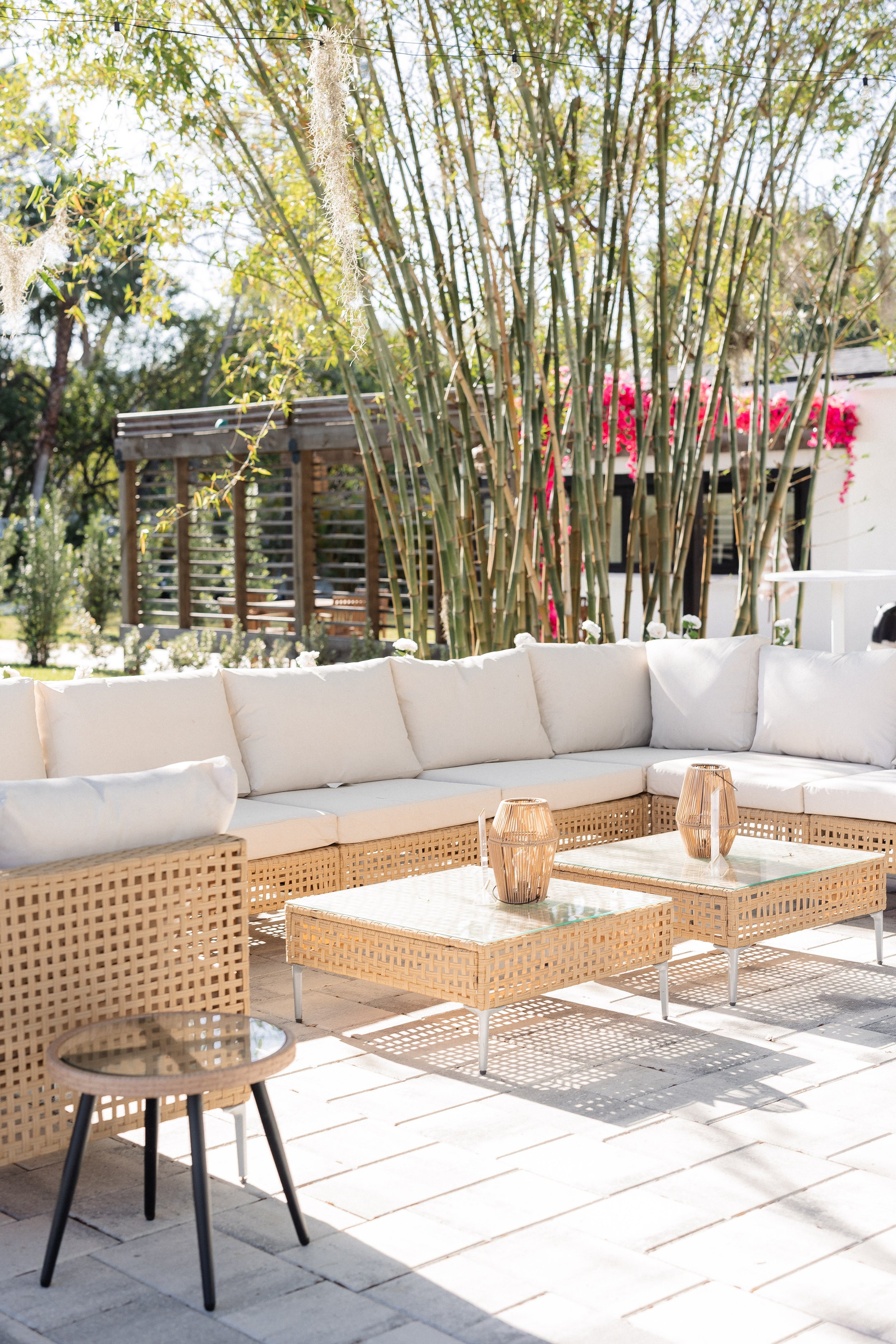 Outdoor patio with beige woven sofa with white cushions, rectangular rattan tables with glass tops and candles, decorative bamboo plants, and a wooden privacy screen in the background, sunny day with trees and pink flowers.