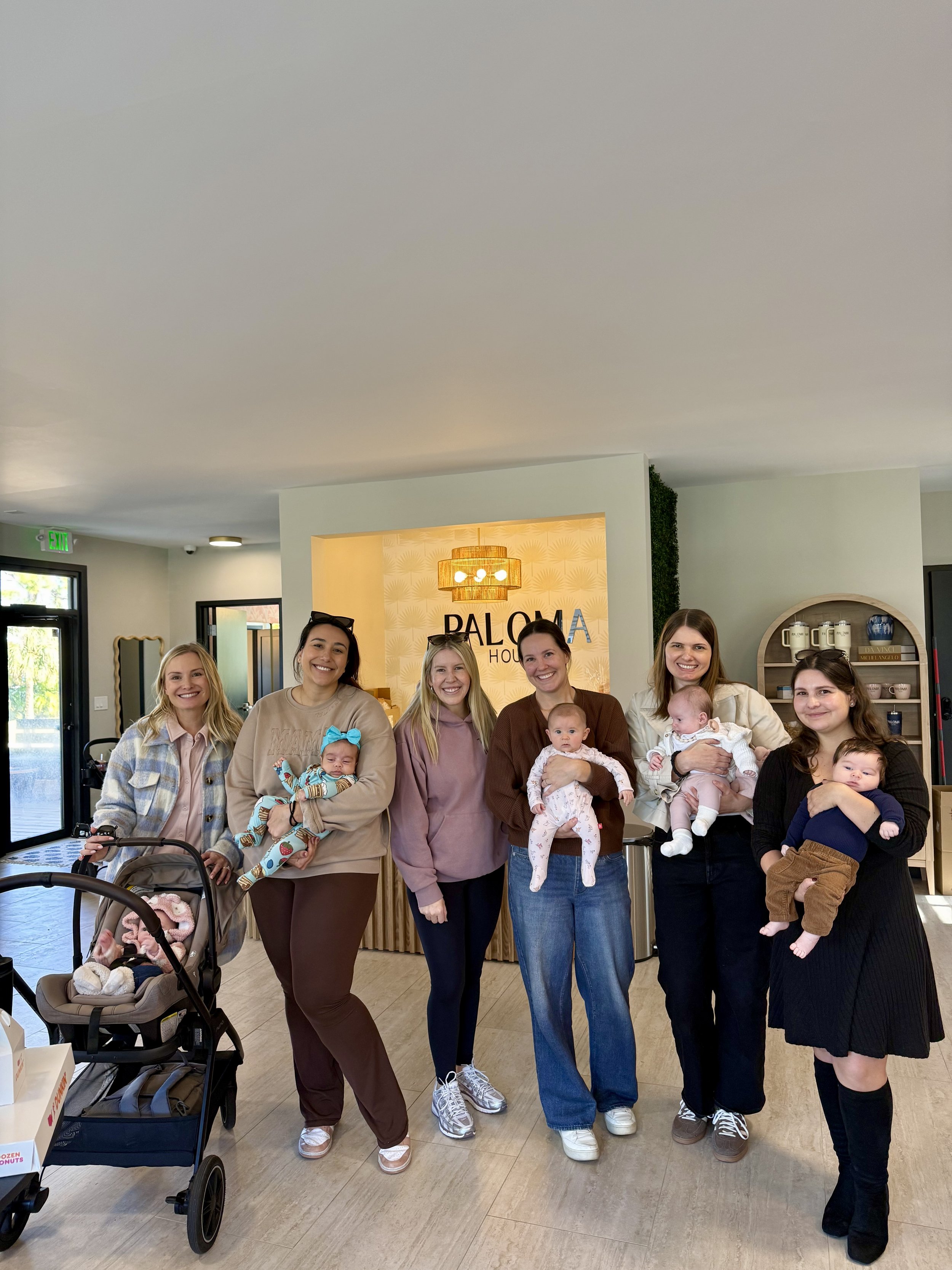 Group of women holding babies inside a modern home or lobby, with a sign that says "BALOMA HO" in the background.