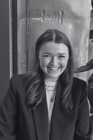 Young woman with long hair smiling, wearing layered necklaces and a blazer, standing in front of a concrete wall.