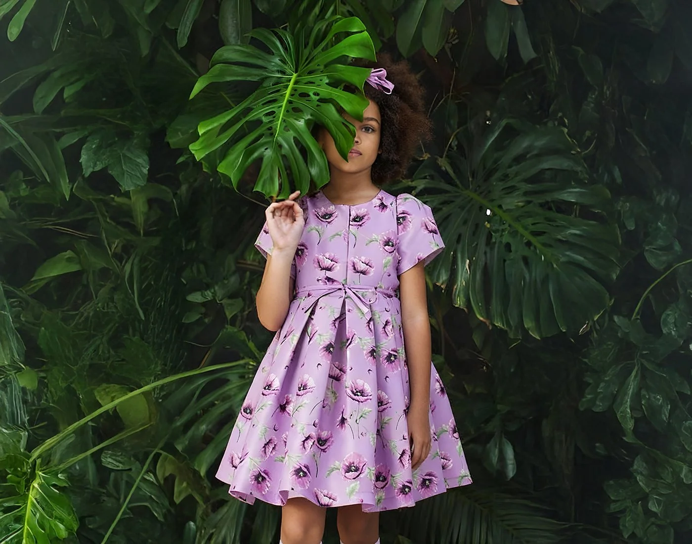 A woman in a purple floral dress standing in front of lush green tropical leaves, holding a large Monstera leaf partially over her face.