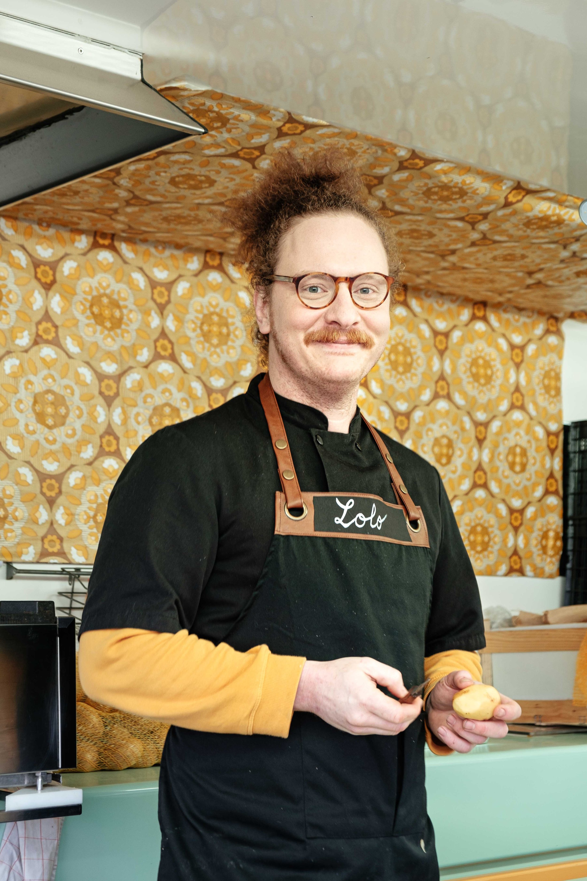 Chef souriant portant un tablier avec le nom 'Lolo' dans une cuisine vintage, tenant une pomme de terre.