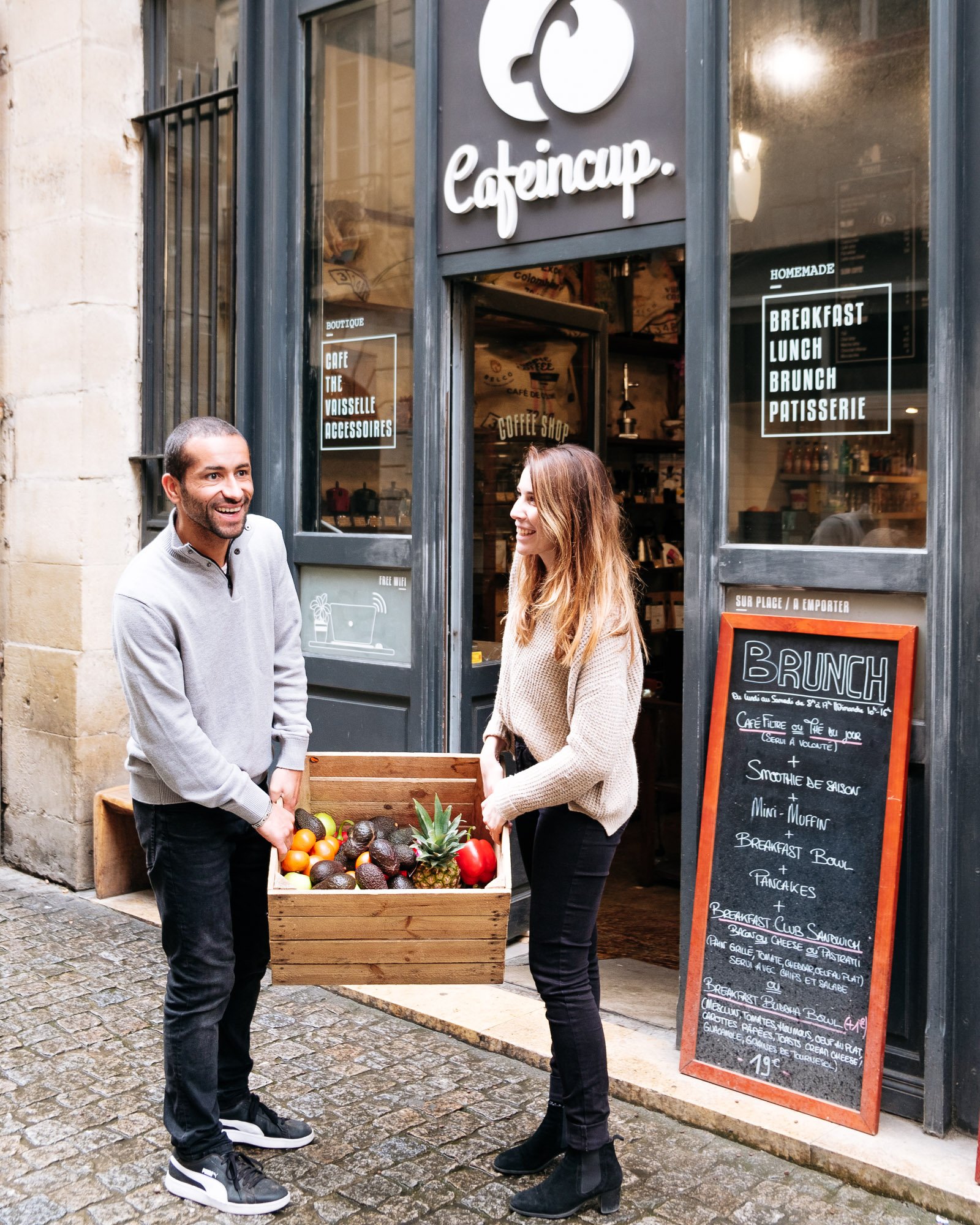 Deux personnes souriantes échangent devant un café avec un panier de fruits exotiques. La façade du café affiche un menu et des inscriptions en français.