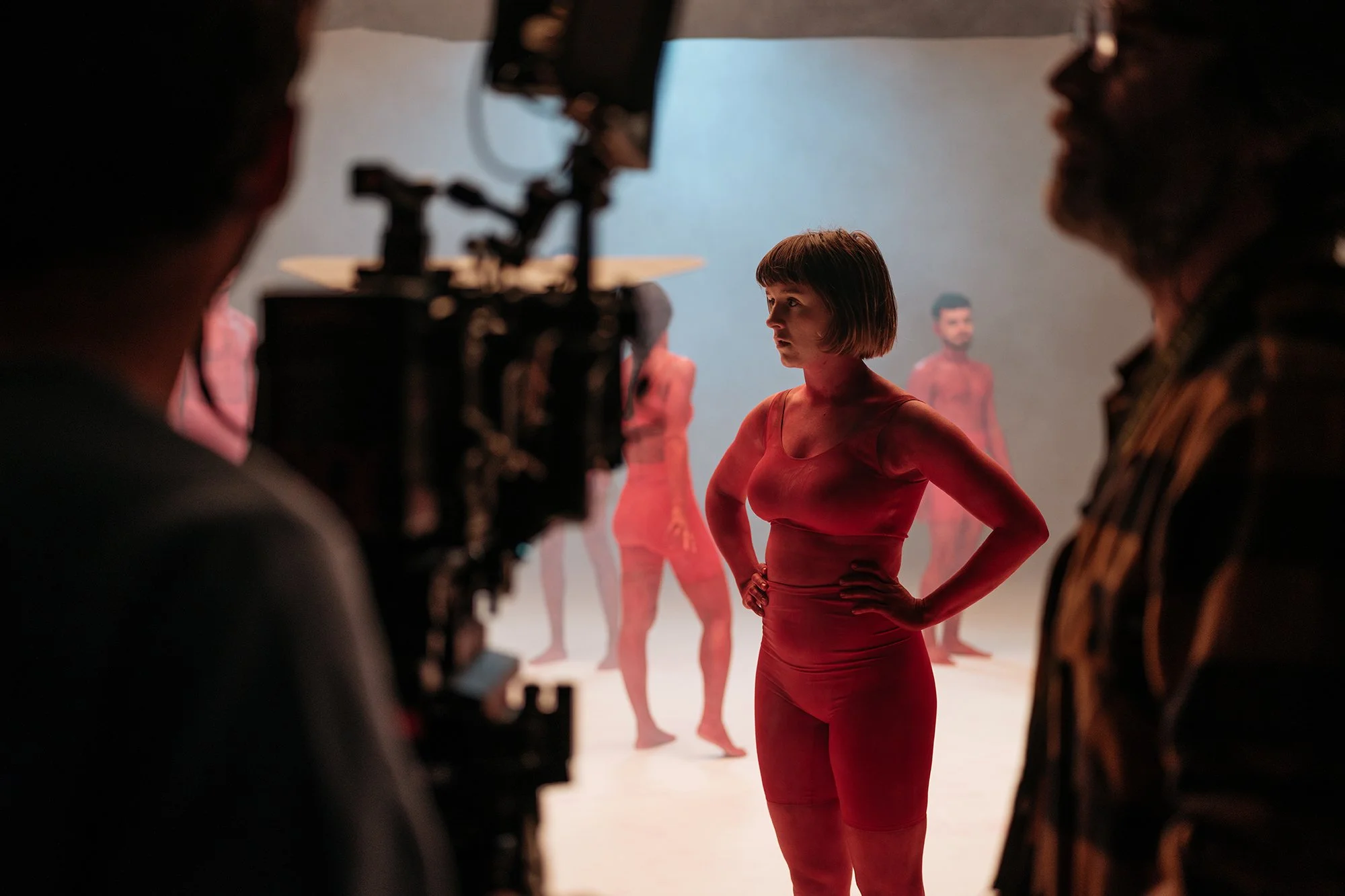 Une femme en costume rouge se tient dans un studio de tournage avec des autres mannequins flous en arrière-plan, entourée de trois membres d'équipe. La scène est sombre avec un éclairage sur la femme.