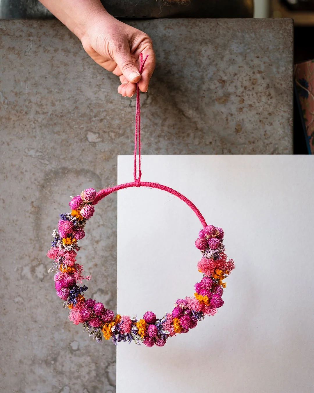Main tenant une couronne de fleurs séchées colorées, principalement roses, avec des touches d'orange, violettes et blanches. La couronne est suspendue par un fil rose, tenue par une main.