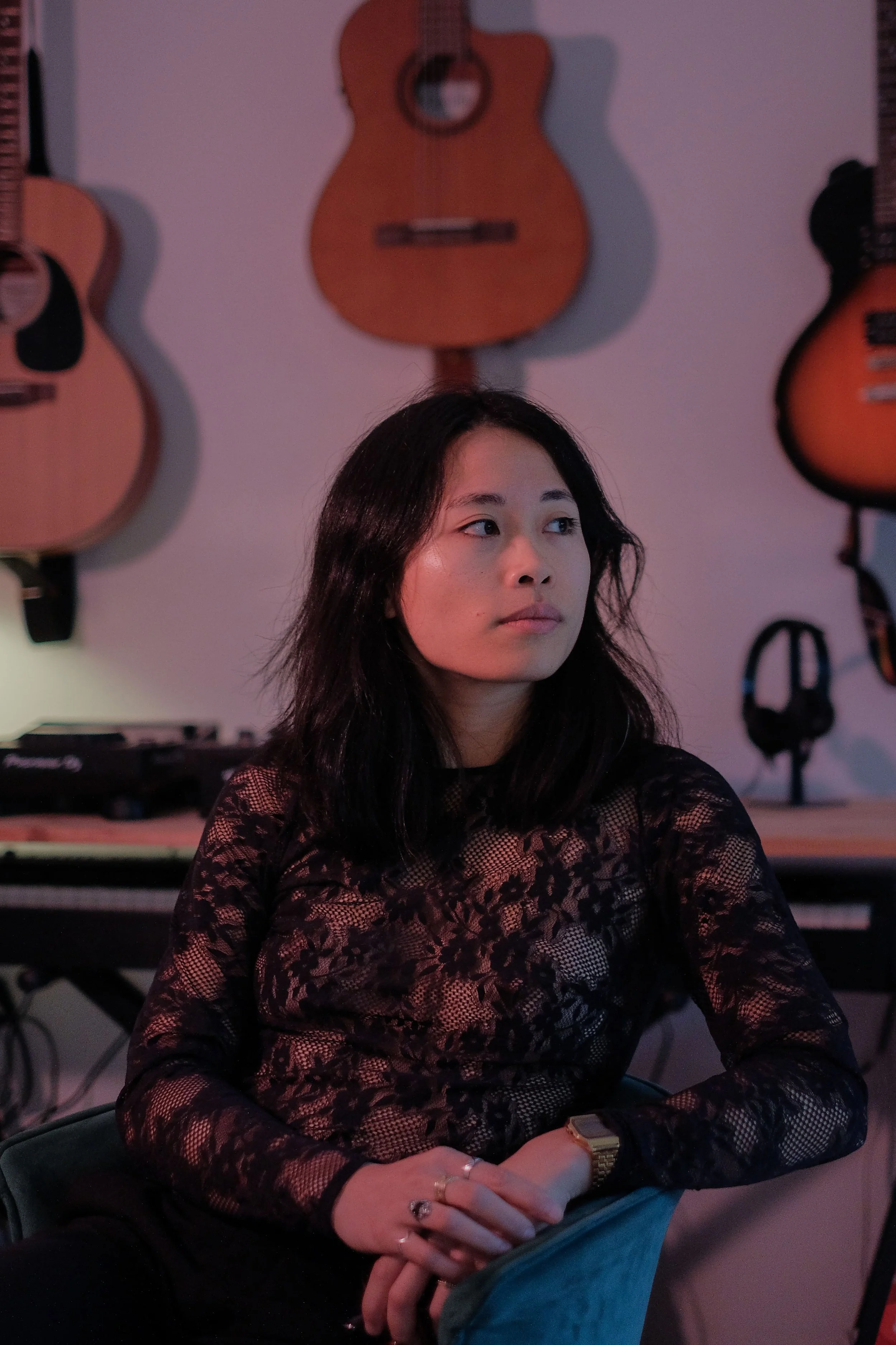 Une femme assise dans une pièce avec des guitares accrochées au mur derrière elle, portant un haut en dentelle noir.