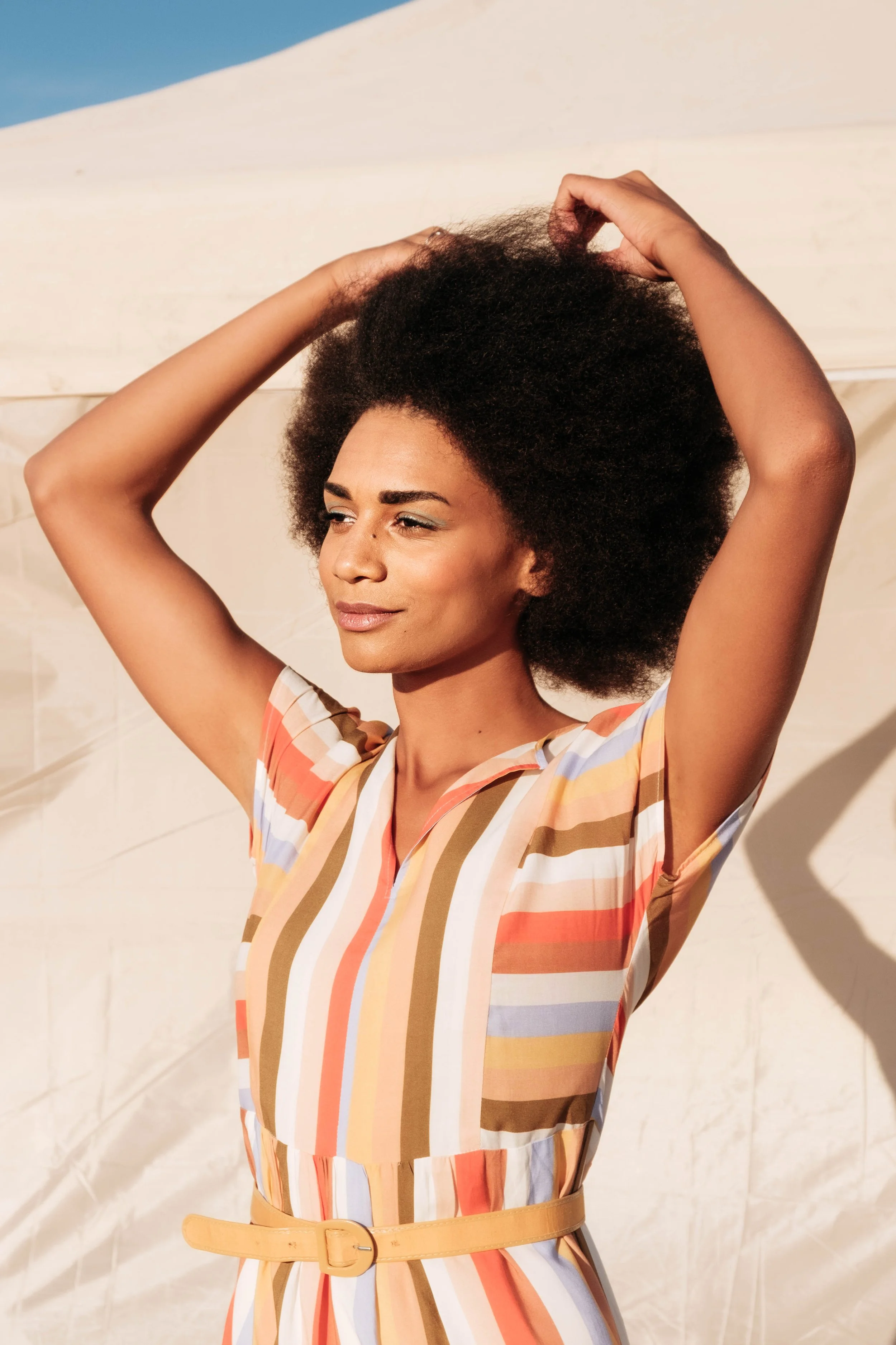 Jeune femme noire avec une coiffure afro, portant une robe à rayures multicolores, en train de profiter du soleil.