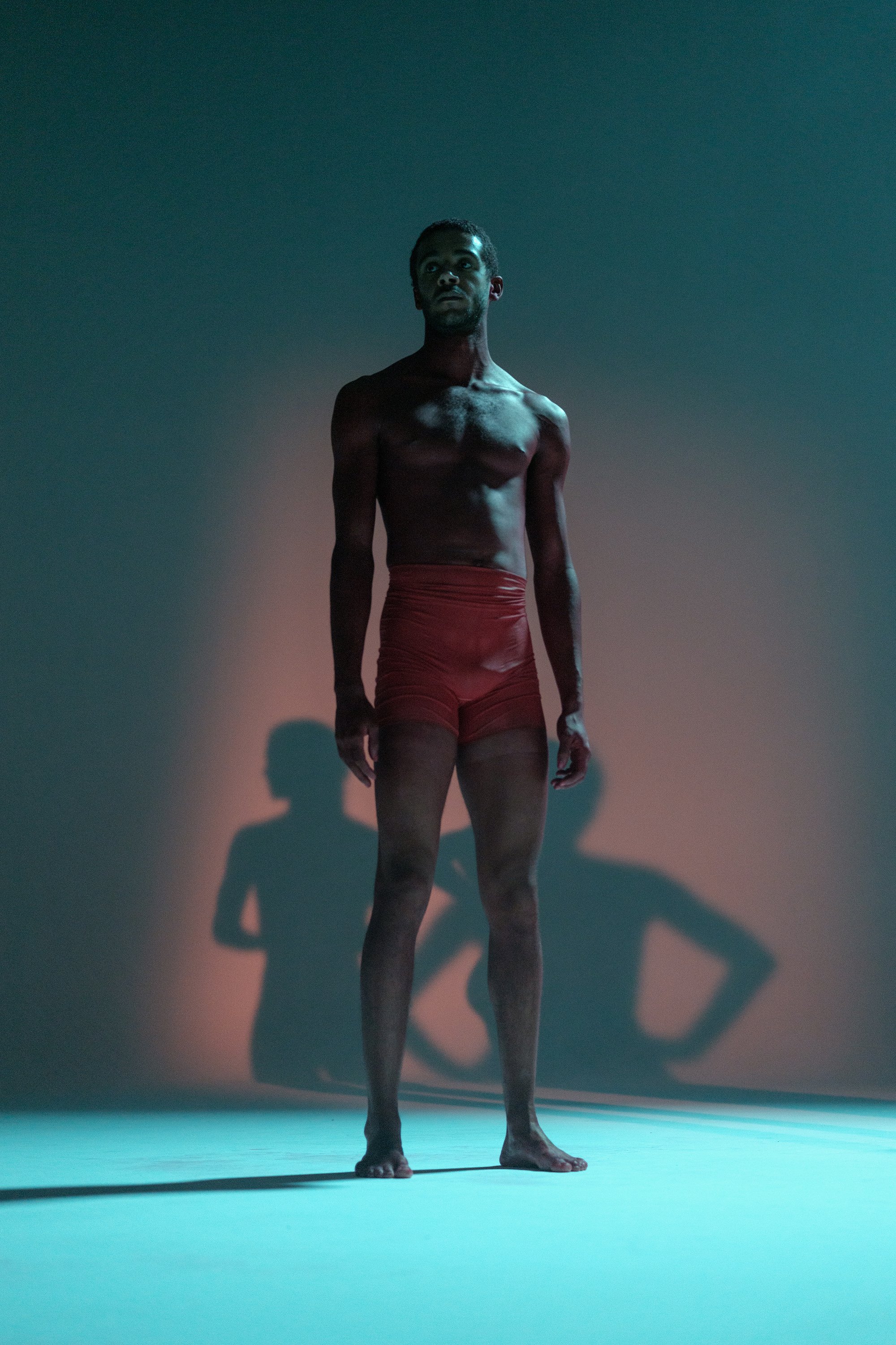 Un homme nu portant un short rouge, debout dans une pièce sombre avec un éclairage coloré, créant une ombre visible sur le mur derrière lui.
