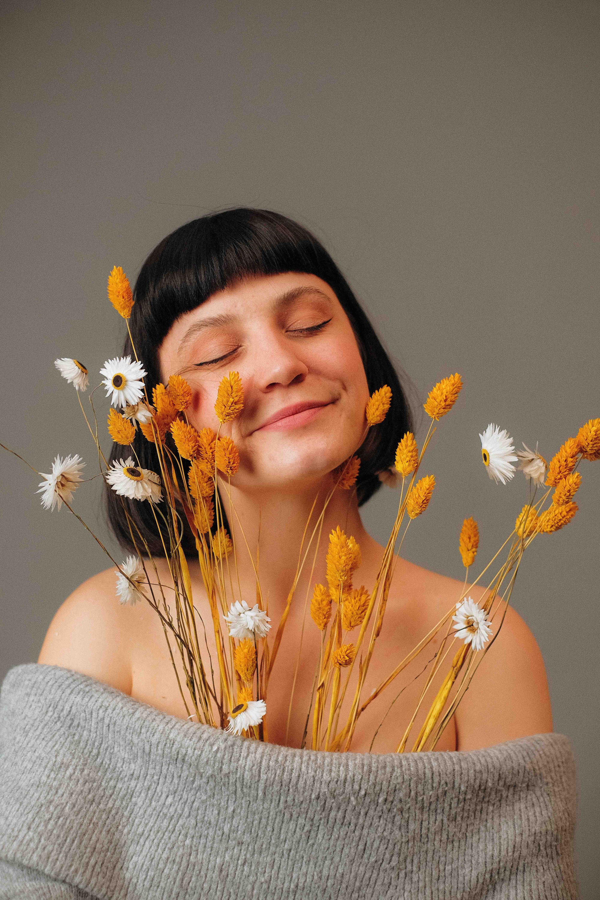 Une femme avec une coiffure courte et noire, ses yeux fermés, tenant un bouquet de fleurs jaunes et blanches, partiellement enveloppée dans un pull gris, sur un fond gris clair.