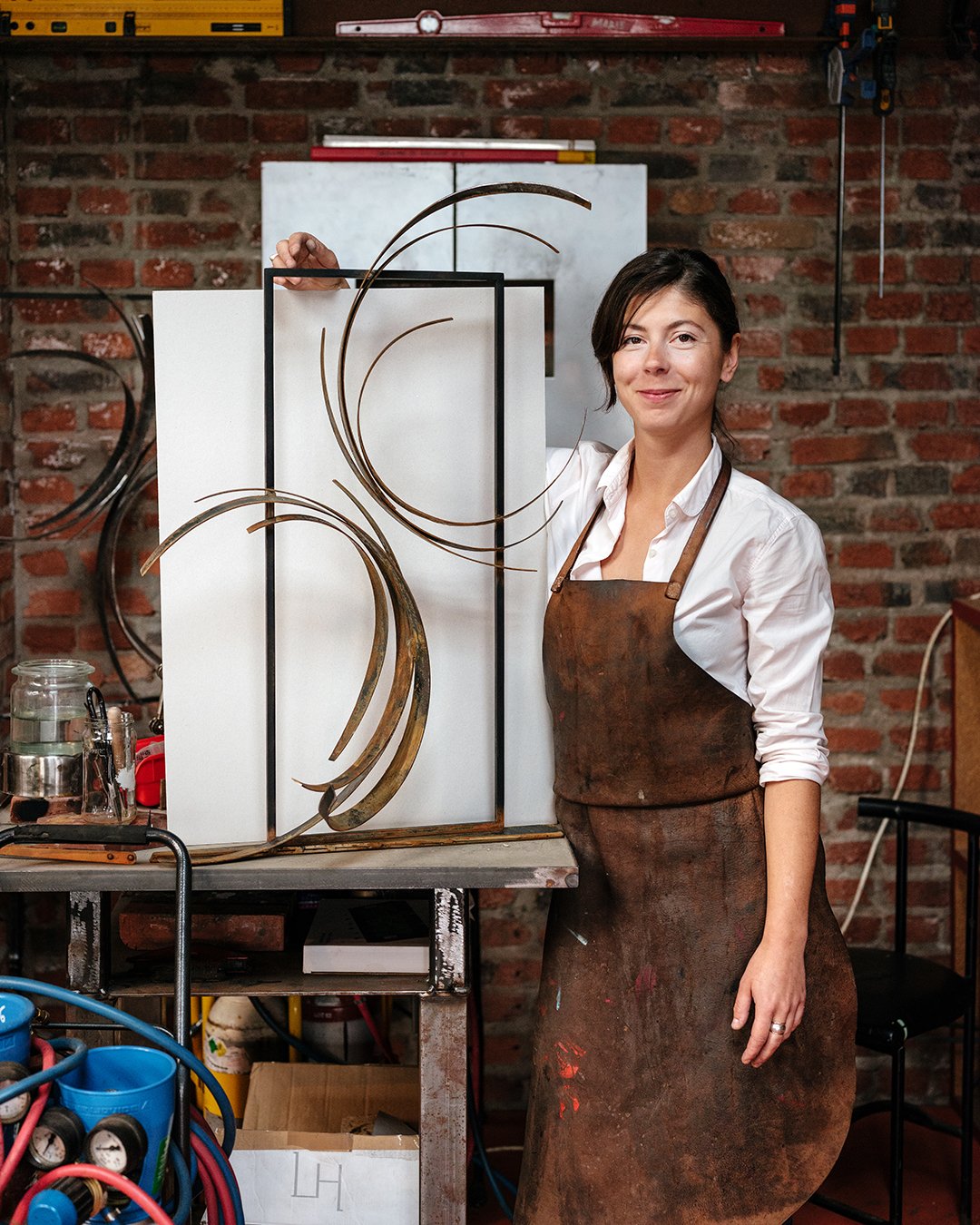 Une femme avec un tablier en cuir rouillé posant dans un atelier avec un fond de mur en briques rouges et diverses fournitures de travail.