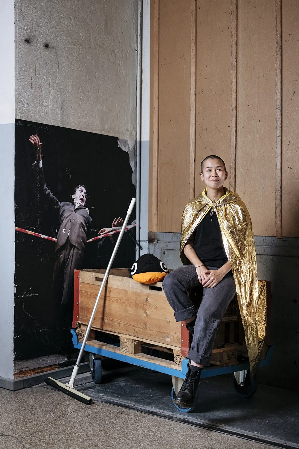 Une jeune femme assise sur une caisse en bois avec une cape dorée. Elle sourit et regarde légèrement vers la droite. À côté d'elle, il y a un sac en forme de personnage avec des yeux et un bec orange. En arrière-plan, une affiche montre une personne 