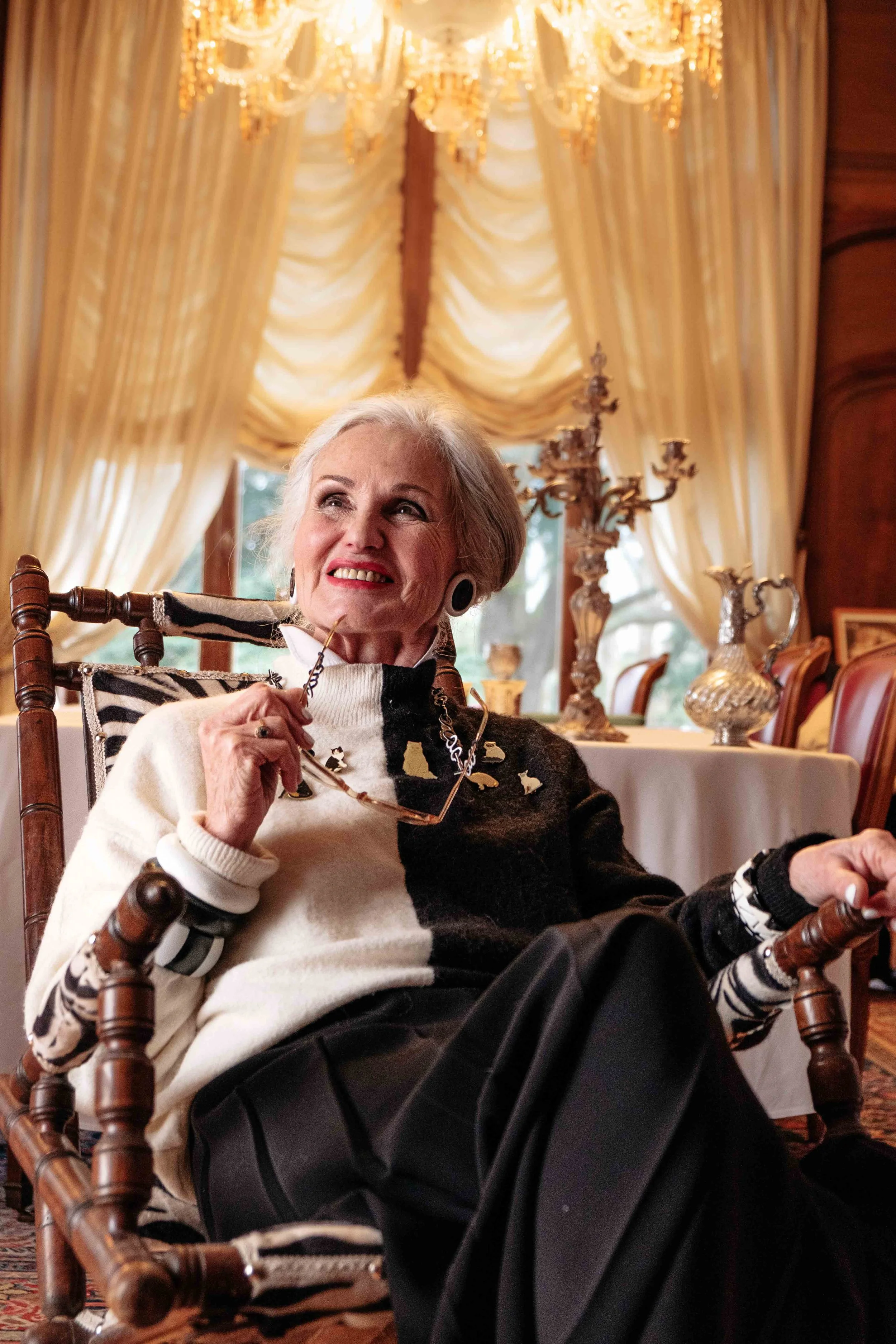 Une femme âgée souriante assise dans une salle élégante avec des rideaux en velours crème et un chandelier chandelier en cristal.