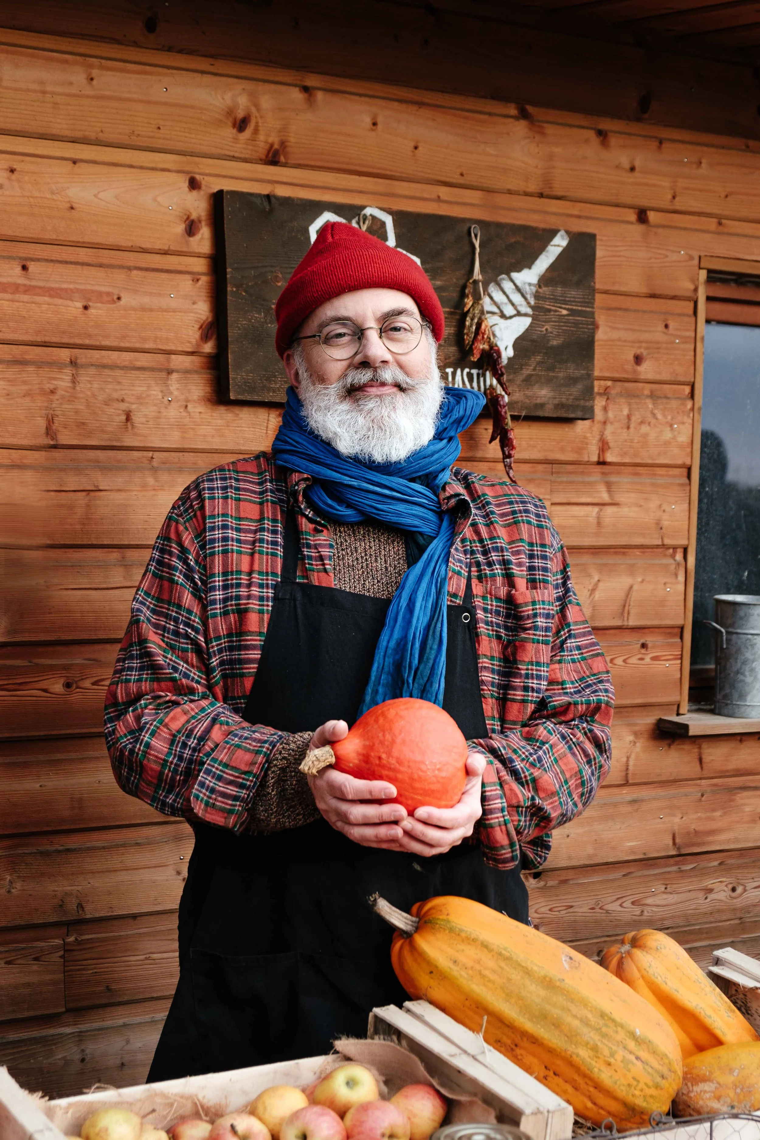 Un homme âgé avec une barbe blanche, portant un bonnet rouge, une écharpe bleue, une chemise à carreaux et un tablier noir, tient une petite citrouille orange. Il est dans un marché de ferme avec un stand en bois, des courges et des pommes sur la tab