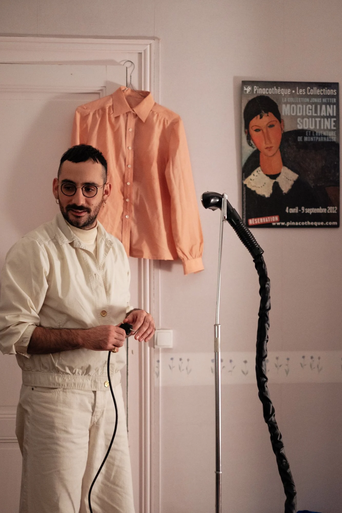 Un homme avec des lunettes, portant une tenue beige, tenant un câble électrique, se tient devant une blouse rose accrochée au mur, à côté d'une affiche de musée dans une pièce intérieure.