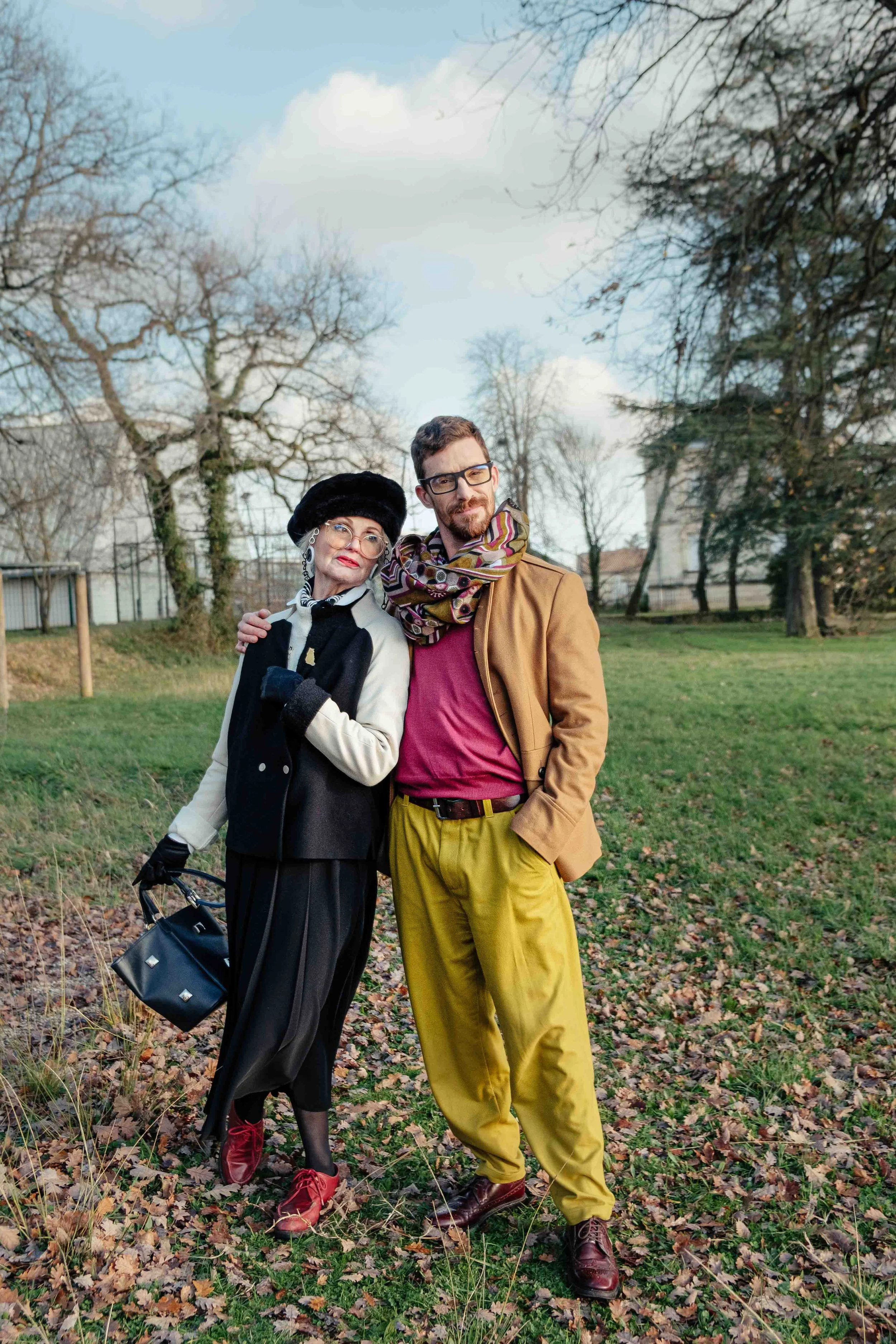 Une femme âgée et un jeune homme posent ensemble dans un parc, souriant. La femme porte un manteau noir et blanc, un chapeau noir, des lunettes, et tient un sac noir. Le jeune homme porte une veste marron, un foulard multicolore, un t-shirt rouge, pa