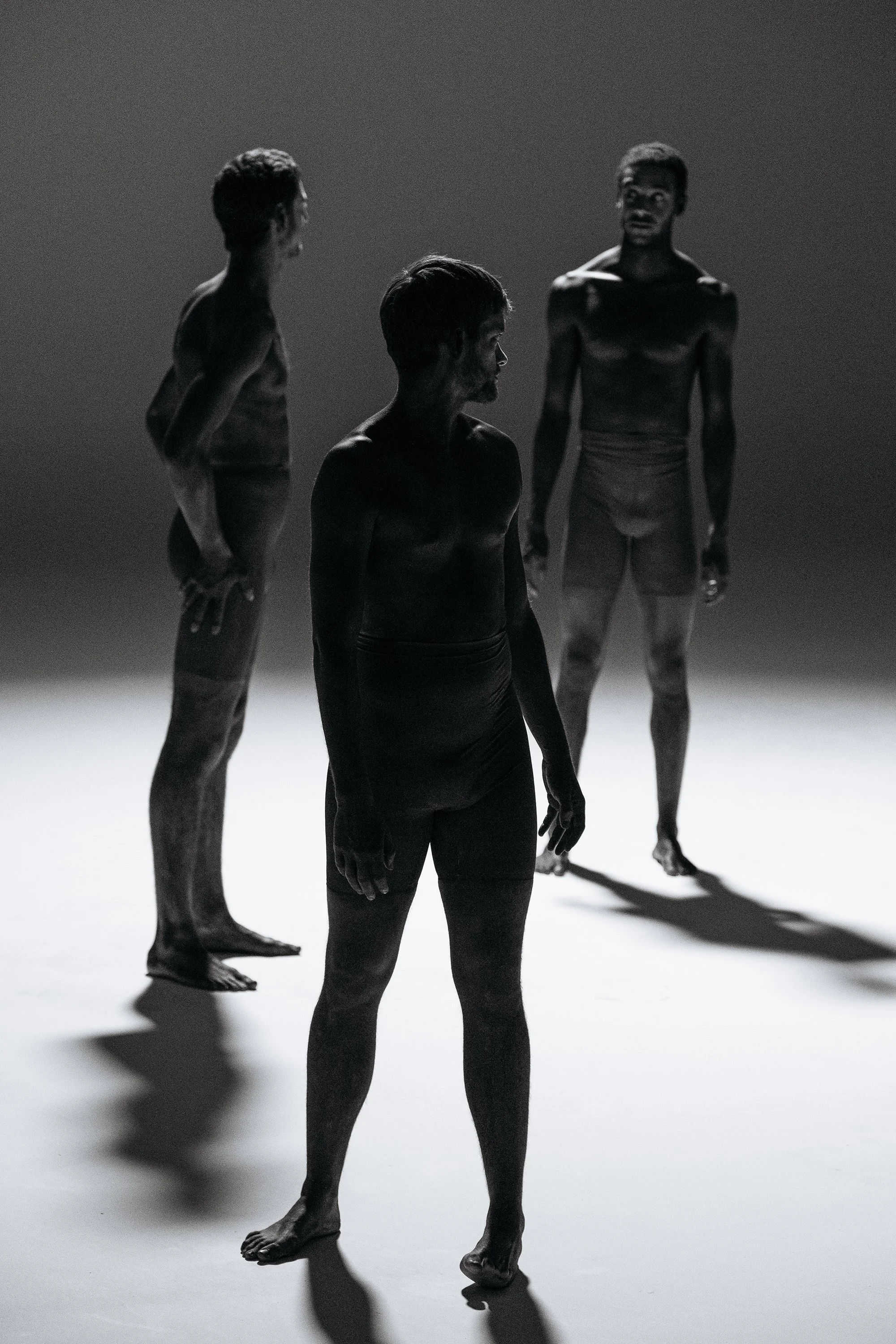 Trois hommes debout, vus de profil, dans un éclairage sombre, créant des silhouettes noires contre un fond gris.