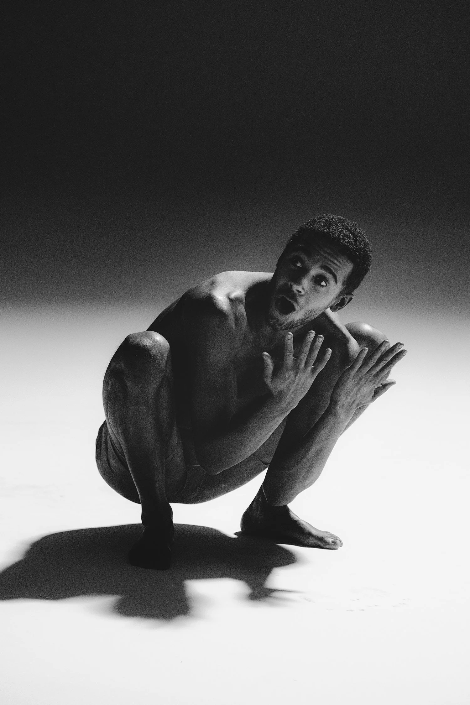 Un homme nu, en position accroupie, avec une expression de surprise ou d'étonnement, en noir et blanc.