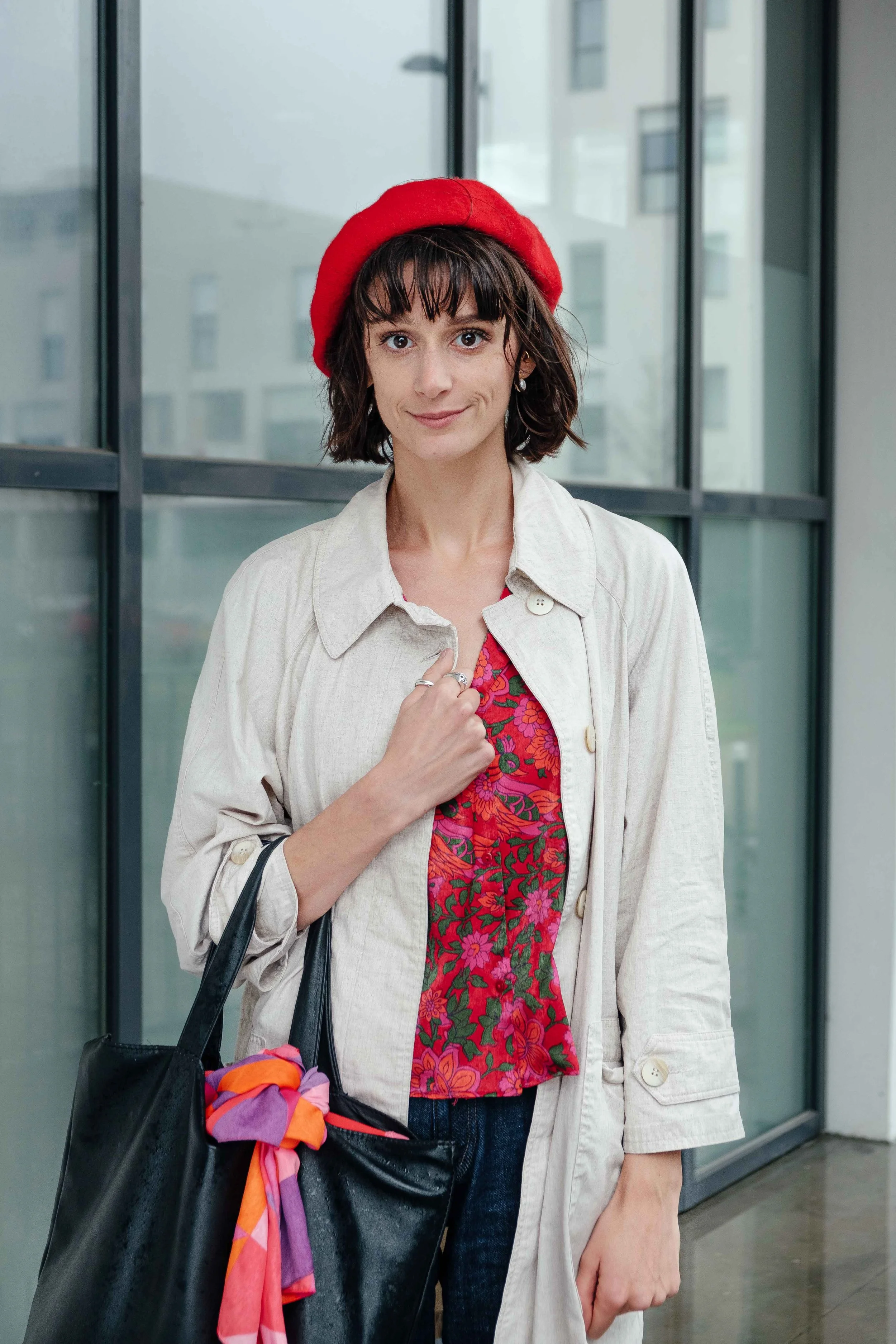 Une femme porte un béret rouge, un manteau beige clair, un chemisier rouge à motifs floraux, et porte un sac noir avec un foulard coloré attaché. Elle se tient dans une pièce avec de grandes fenêtres.