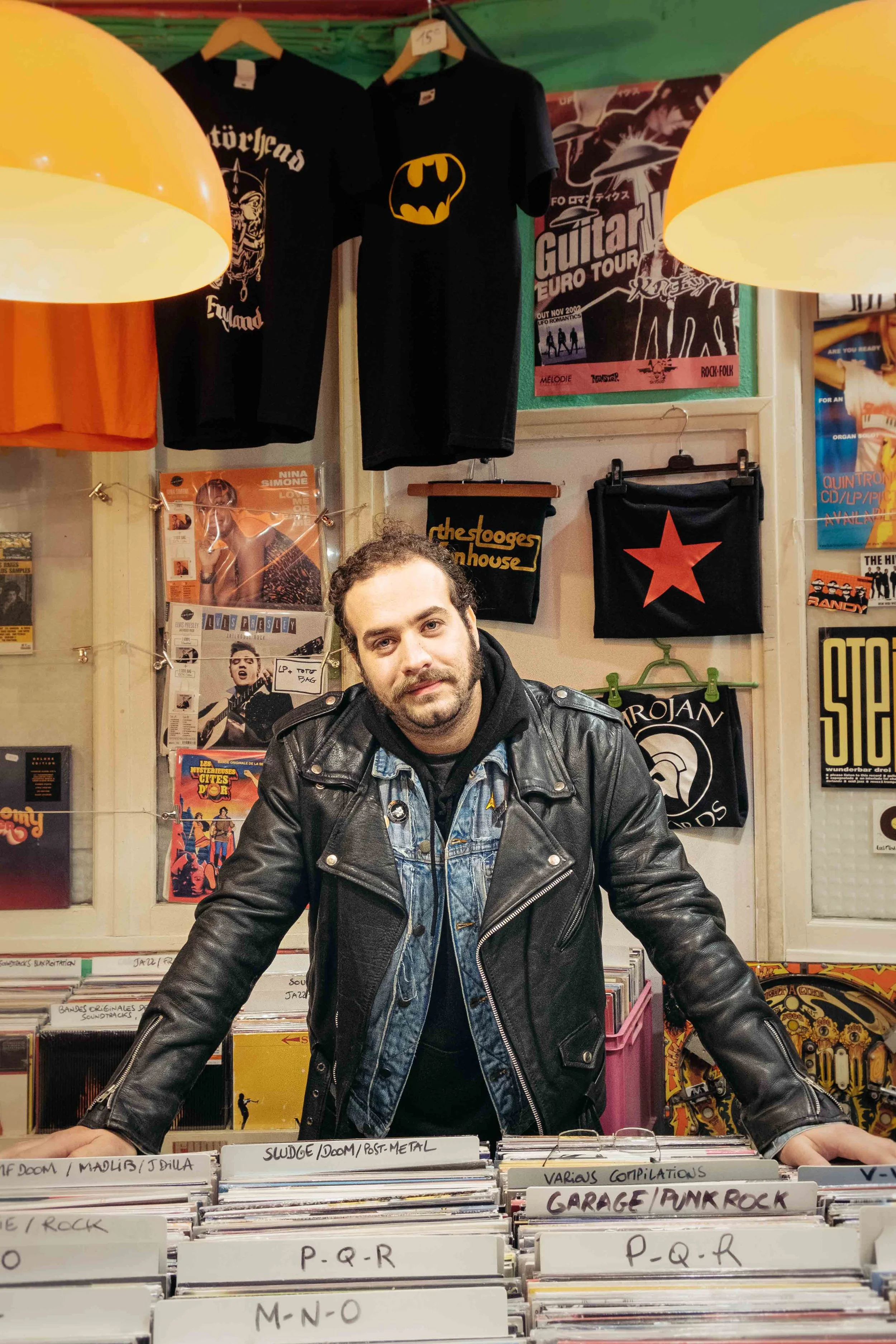 Un homme debout derrière une étagère de disques vinyles dans une boutique de musique, portant une veste en cuir et une chemise en jean, avec des T-shirts à motifs et des affiches au mur derrière lui.