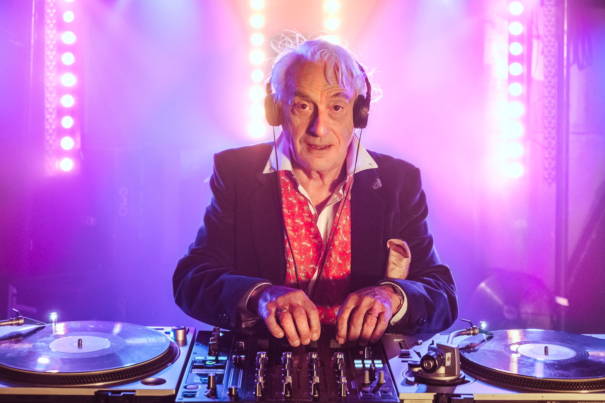 Un homme âgé avec des cheveux gris, portant un casque audio, derrière un set de platines de DJ, éclairé par des lumières violettes et roses.