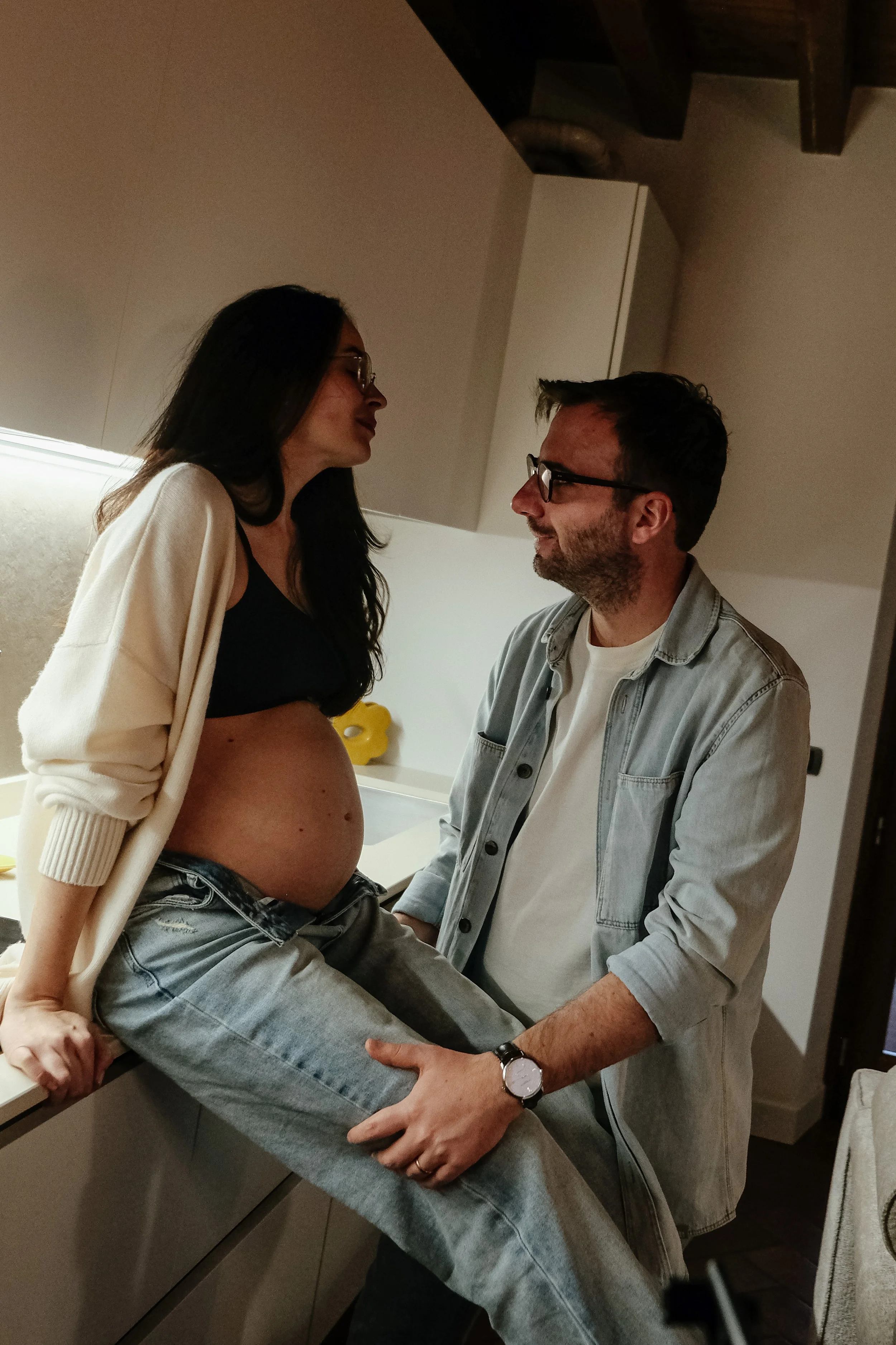Une femme enceinte assise sur un comptoir de cuisine, face à un homme qui lui tient la main, dans une cuisine moderne.