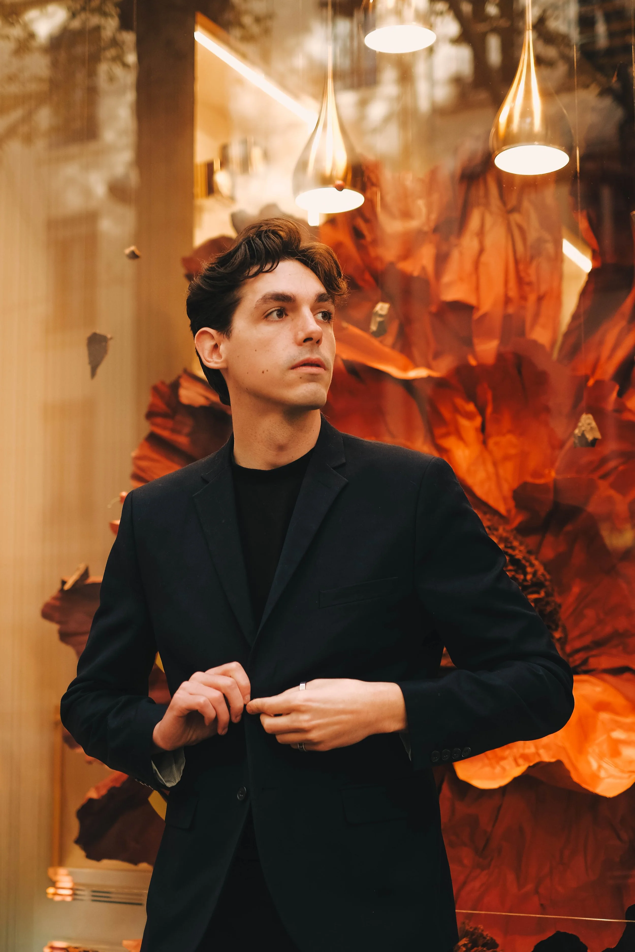 Un jeune homme en costume noir, debout devant une sculpture en papier orange dans une vitrine avec des lampes suspendues au plafond.