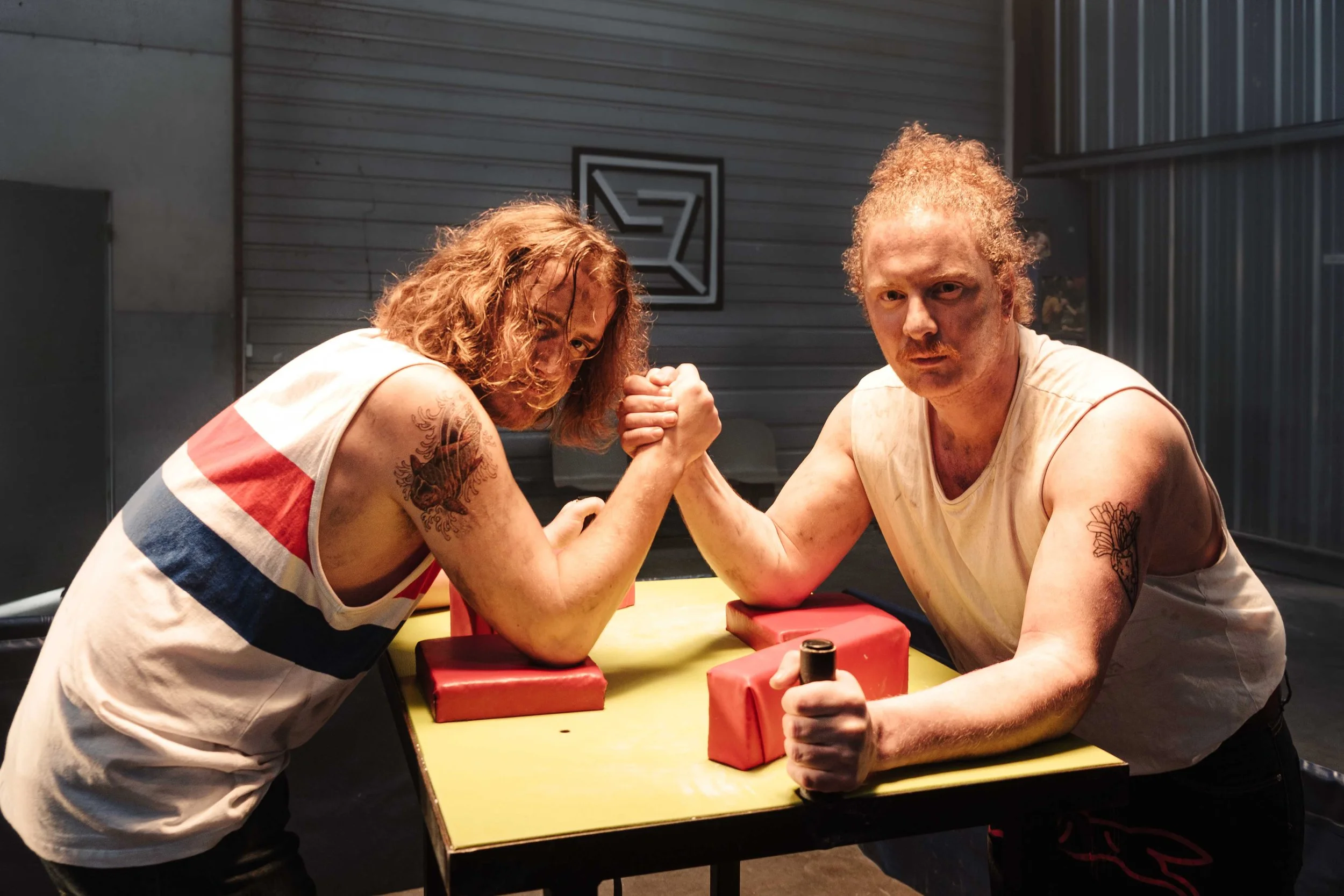 Deux hommes engagés dans une compétition de bras de fer sur une table, chacun tenant la main de l'autre avec intensité. L'arrière-plan est industriel avec un mur en métal et un logo noir et blanc.