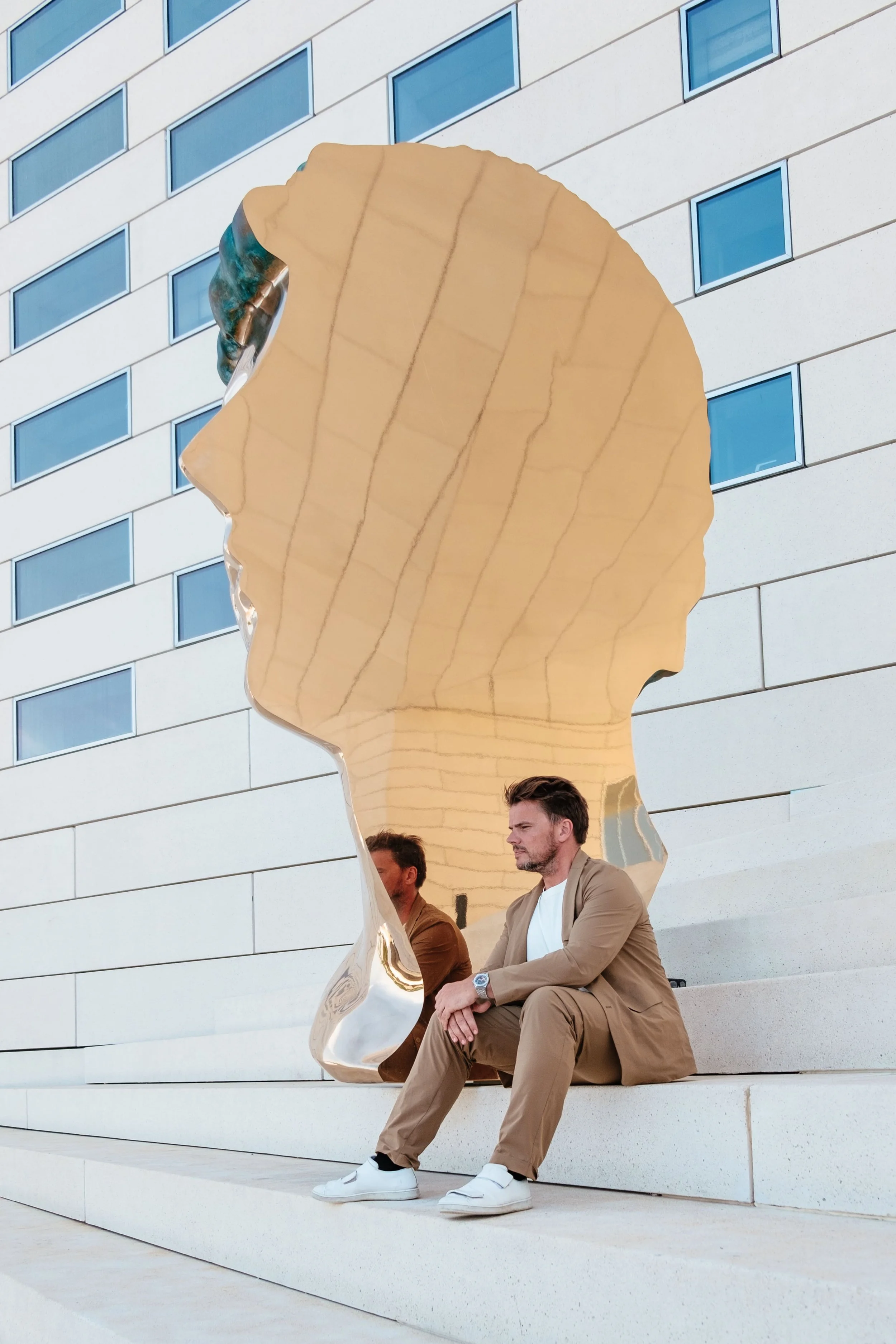 Bjarke Ingels devant MECA à Bordeaux. Assis sur un banc devant une sculpture en forme de profil d'une tête humaine en métal miroir, avec un bâtiment moderne en arrière-plan.