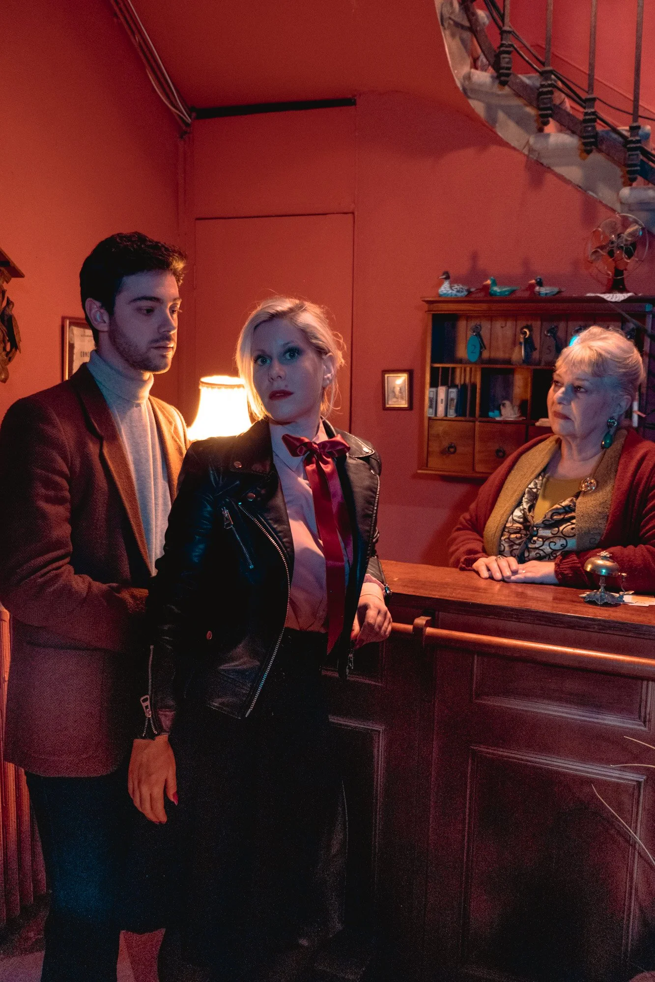 Deux jeunes adultes, un homme et une femme, se tiennent près d'un comptoir dans un intérieur au décor vintage, tandis qu'une femme âgée est derrière le comptoir, dans une pièce aux murs rouges et à la décoration rétro.