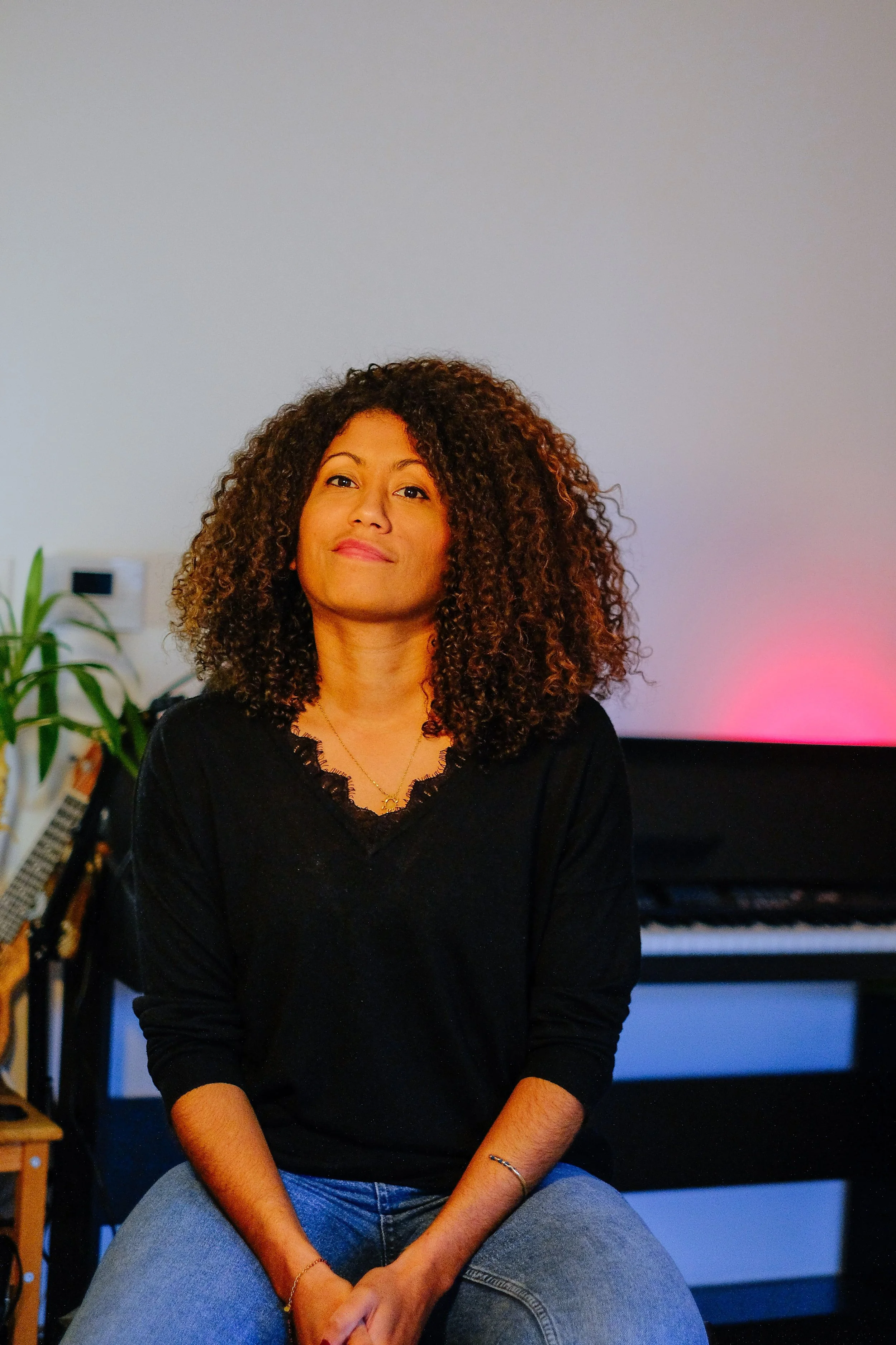 Une femme avec de longs cheveux bouclés assise dans une pièce, avec un piano en arrière-plan et un éclairage coloré rose et bleu.