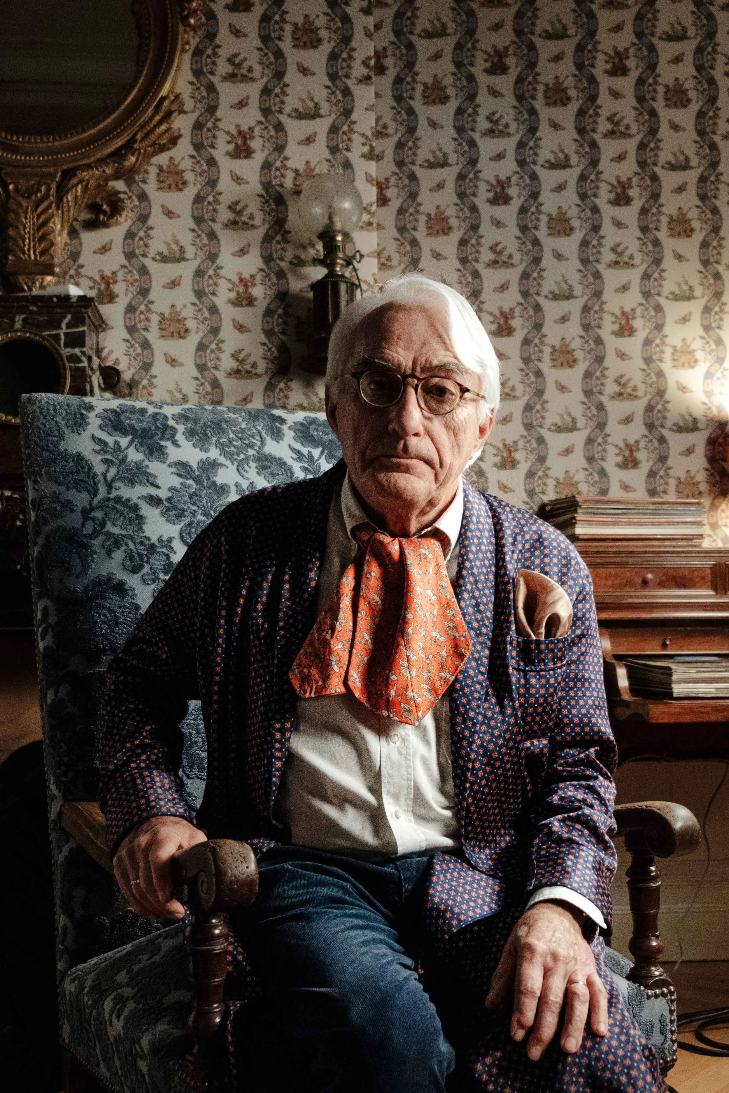 Un homme âgé avec des cheveux blancs, portant des lunettes, une chemise blanche, une veste à motifs et un foulard orange. Il est assis dans un fauteuil dans une pièce décorée avec un papier peint floral, un miroir ancien, une lampe et une pile de mag
