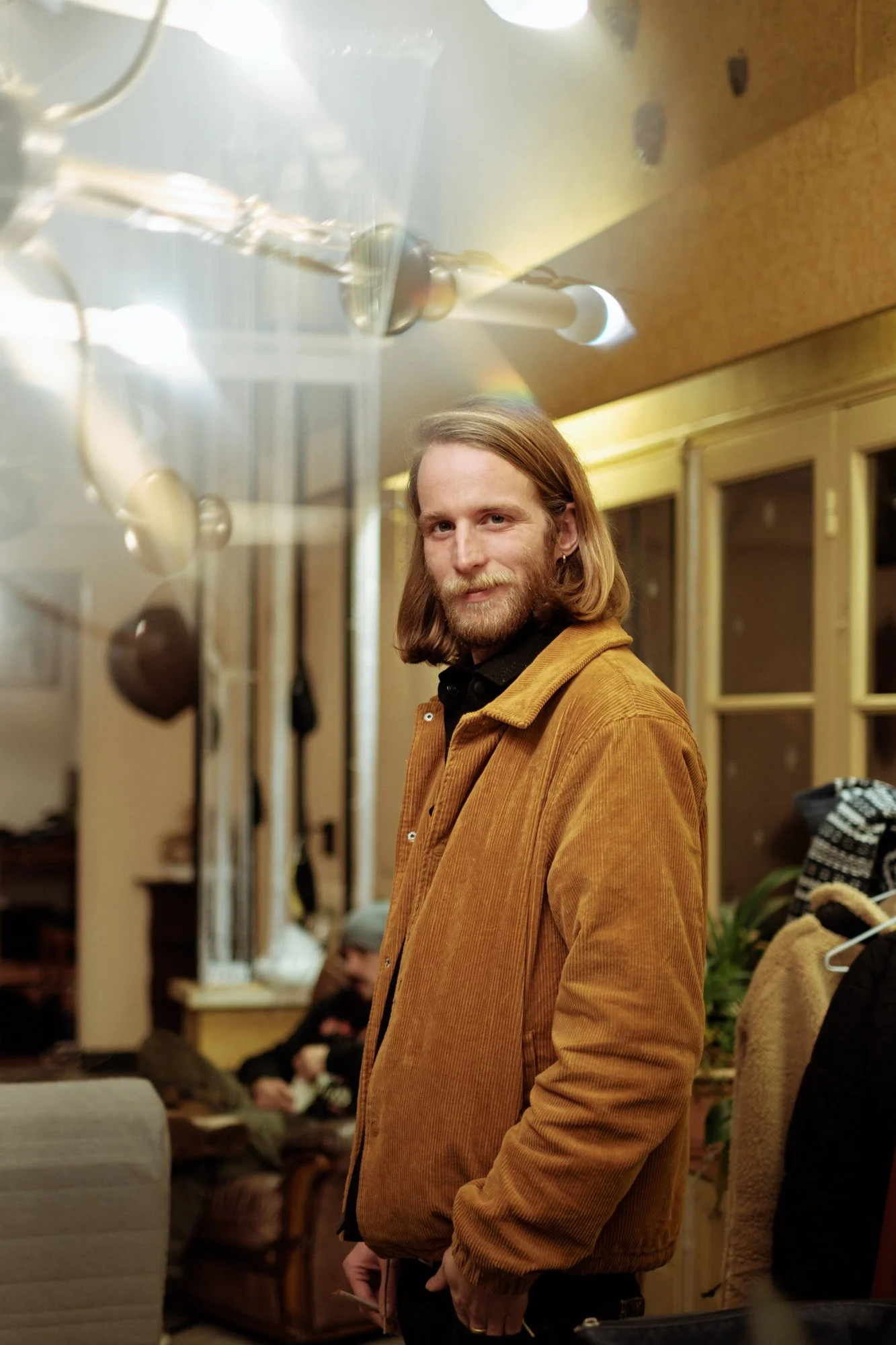 Un homme avec des cheveux longs et une barbe, portant une veste marron, sourit en regardant l'appareil photo dans un intérieur chaleureux, avec des personnes assises en arrière-plan.