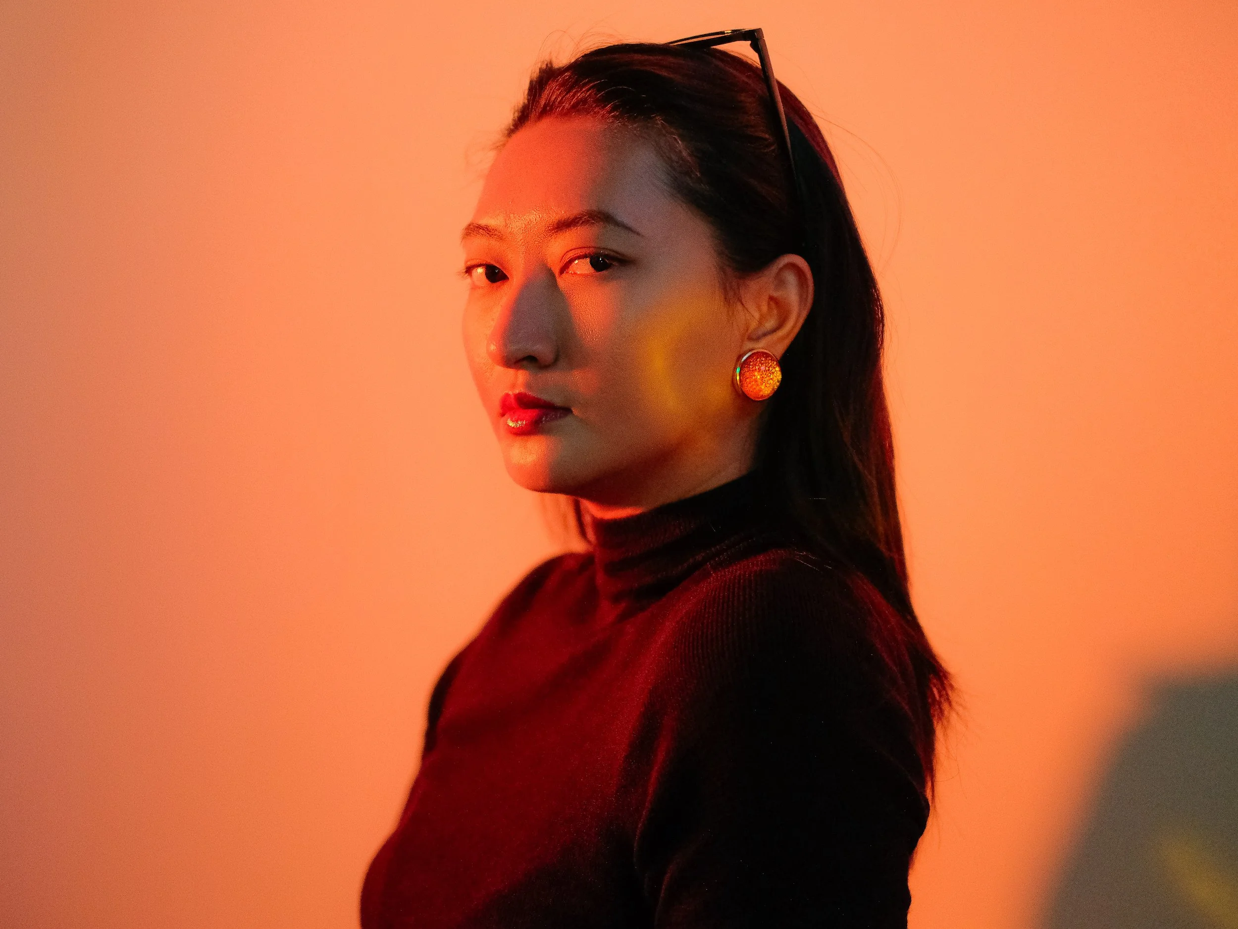 Portrait d'une femme asiatique portrait en gros plan, portant des boucles d'oreilles orange et un haut noir, avec un fond orange et rouge chaud.