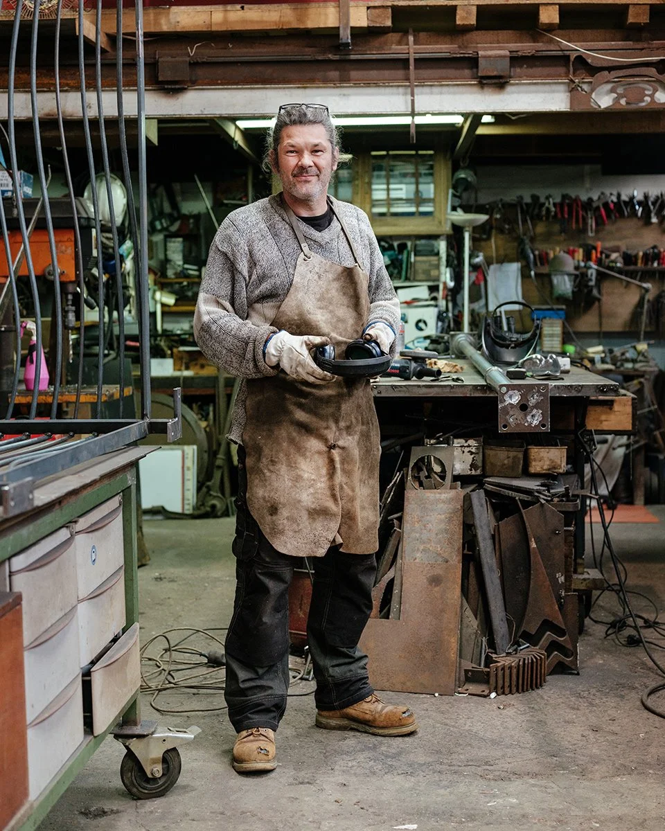 Un homme dans un atelier de bricolage ou de fabrication, portant un tablier en cuir et des gants, tenant un casque de protecteur, entouré d'outils et de matériel suspendu ou posé sur un établi.