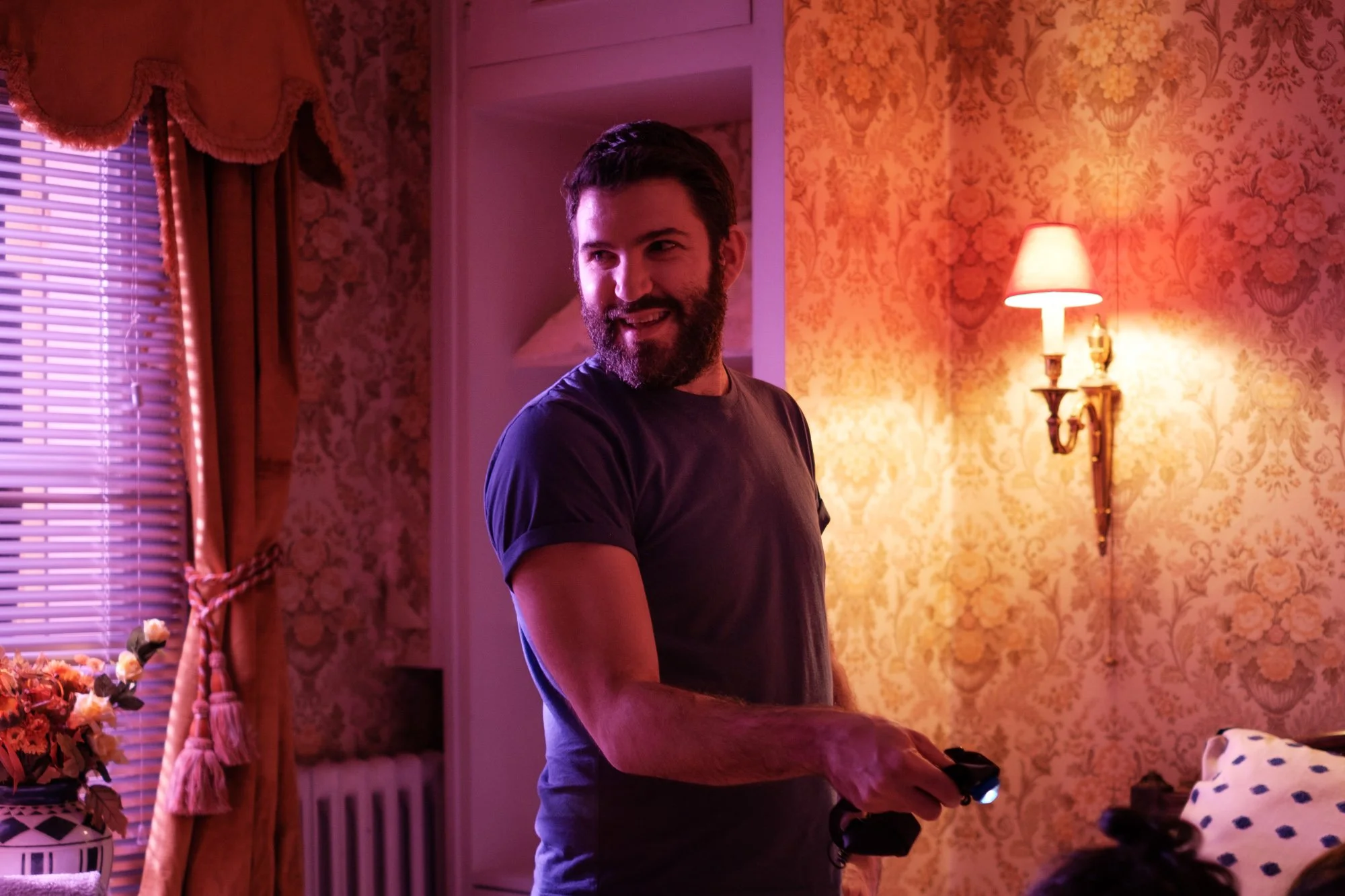 Un homme souriant avec une barbe, dans une pièce au décor vintage avec des papiers peints floraux, tient une manette de jeu vidéo.
