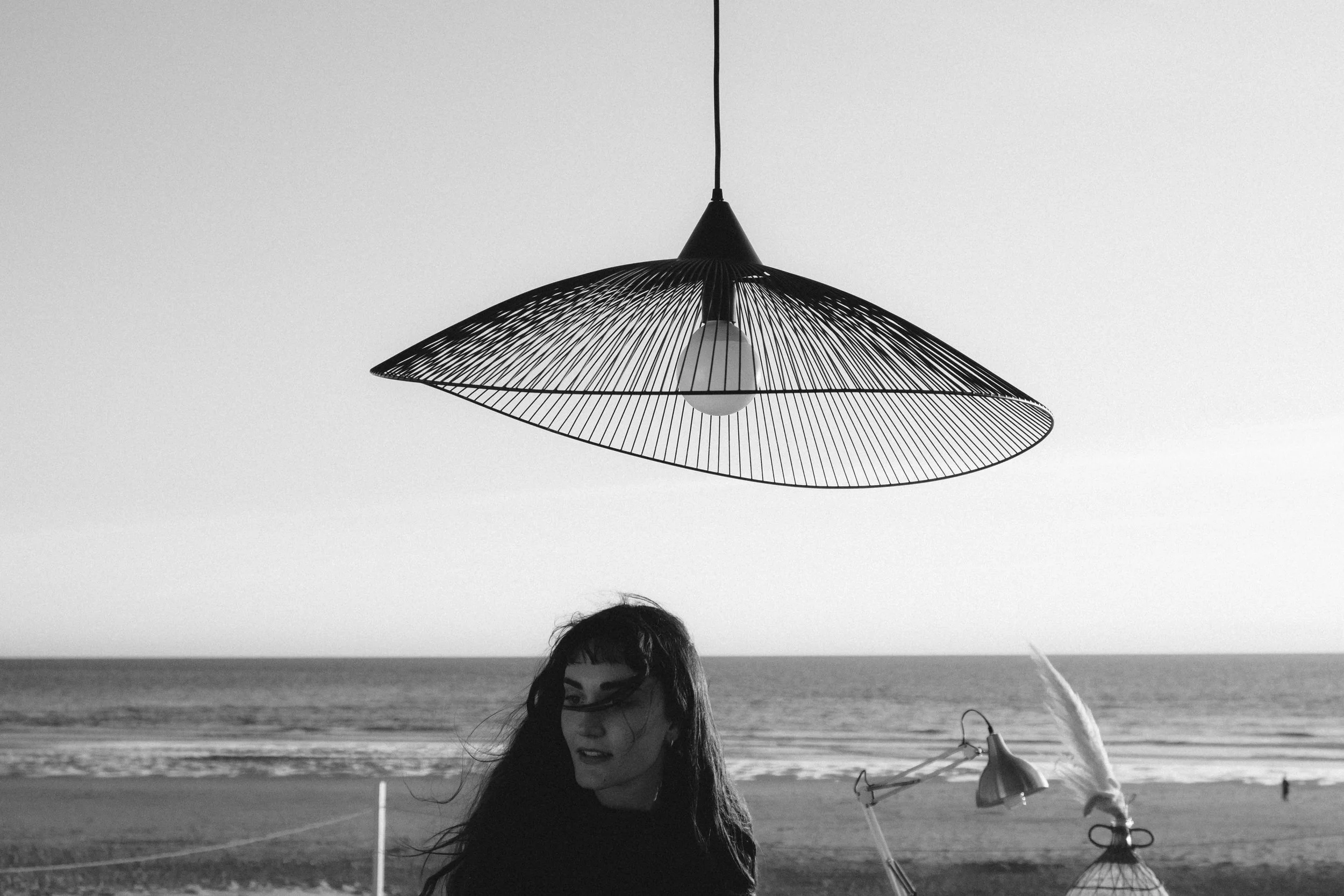 Une femme avec des cheveux longs et bouclés, portant des lunettes noires, se tient sur une plage près de l'océan, avec un parasol noir en forme de disque suspendu au-dessus d'elle. La scène est en noir et blanc.