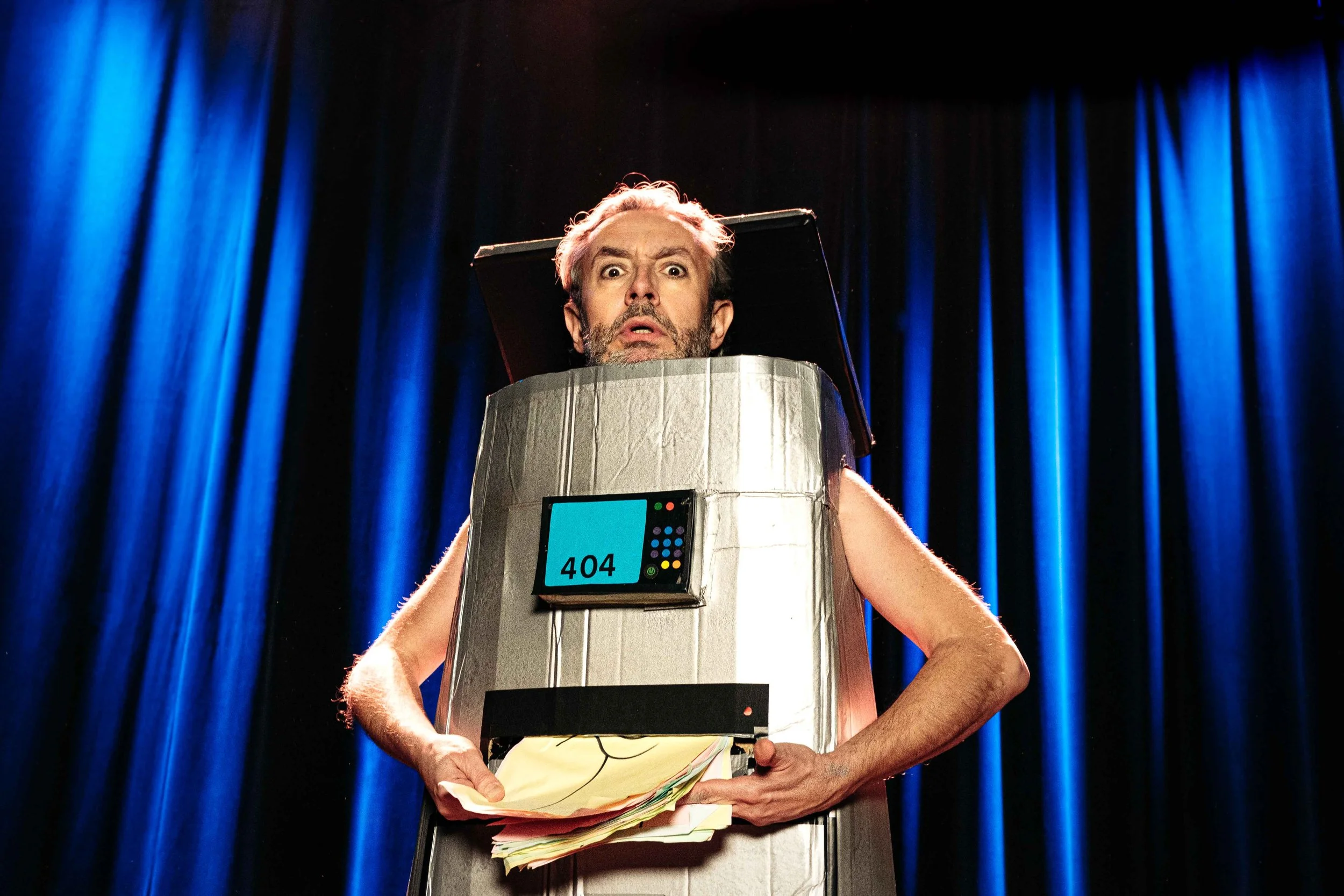 Un homme habillé d'un costume de robot en papier aluminium, avec un écran affichant '404', se tenant sur scène avec un fond de rideaux bleus.
