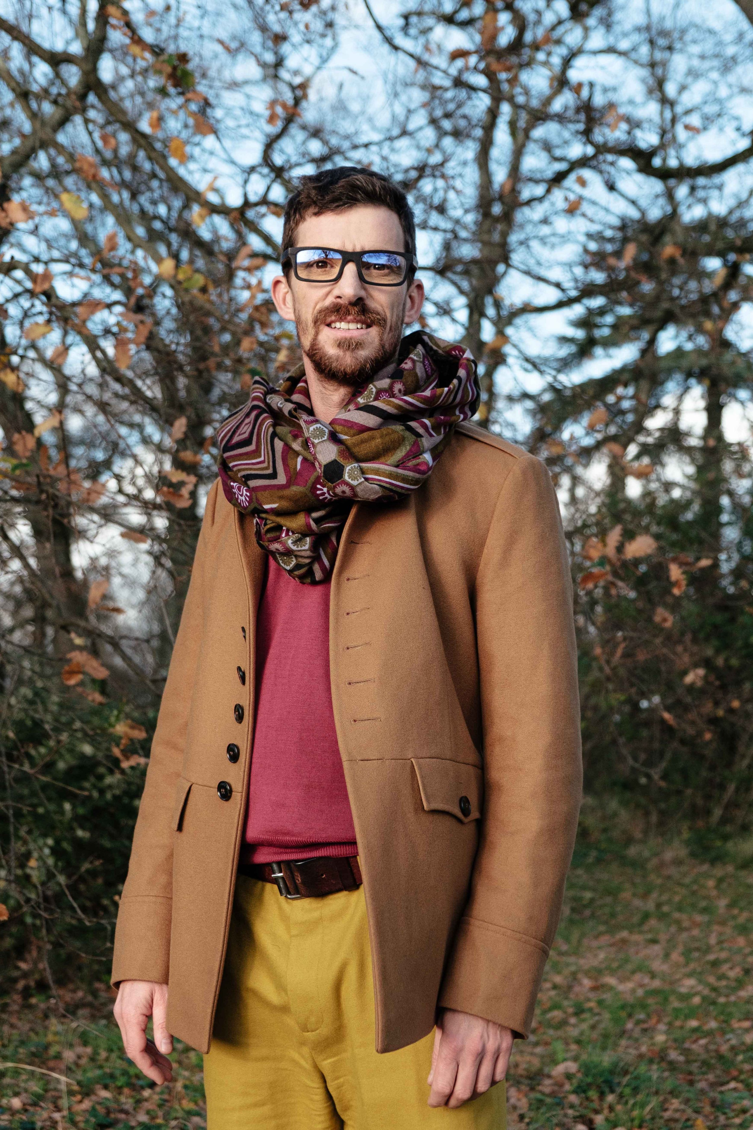 Un homme posant à l'extérieur parmi des arbres, portant un manteau marron, une écharpe, des lunettes et un pull rouge, avec un paysage automnal en arrière-plan.